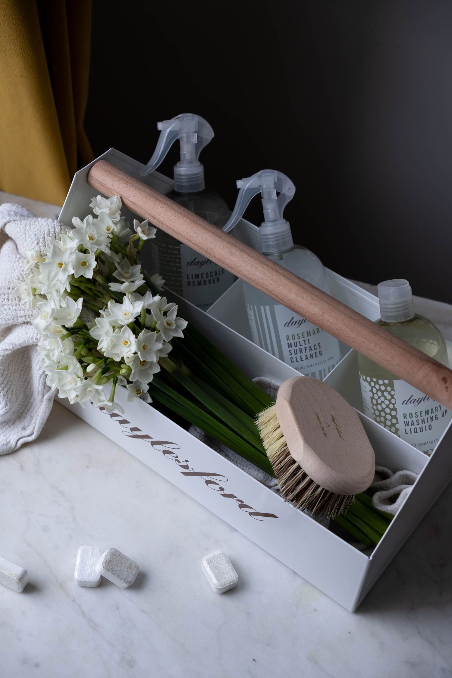 Daylesford cleaning caddy with spray bottles, natural brush, white flowers, and folded cloth on marble surface.