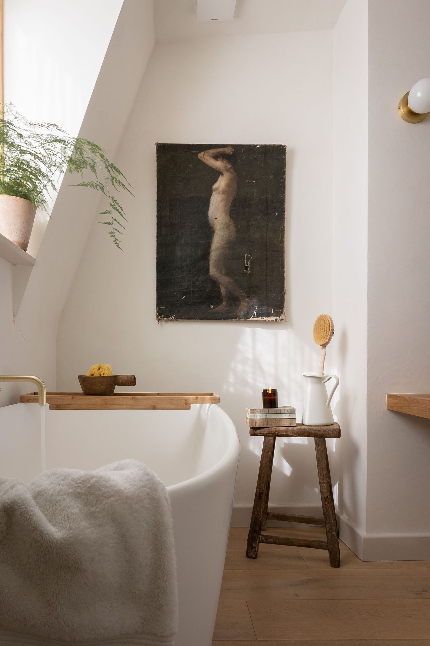Minimalist white bathroom with freestanding tub, wooden stool, brass accents, dark figurative artwork, and green plant.