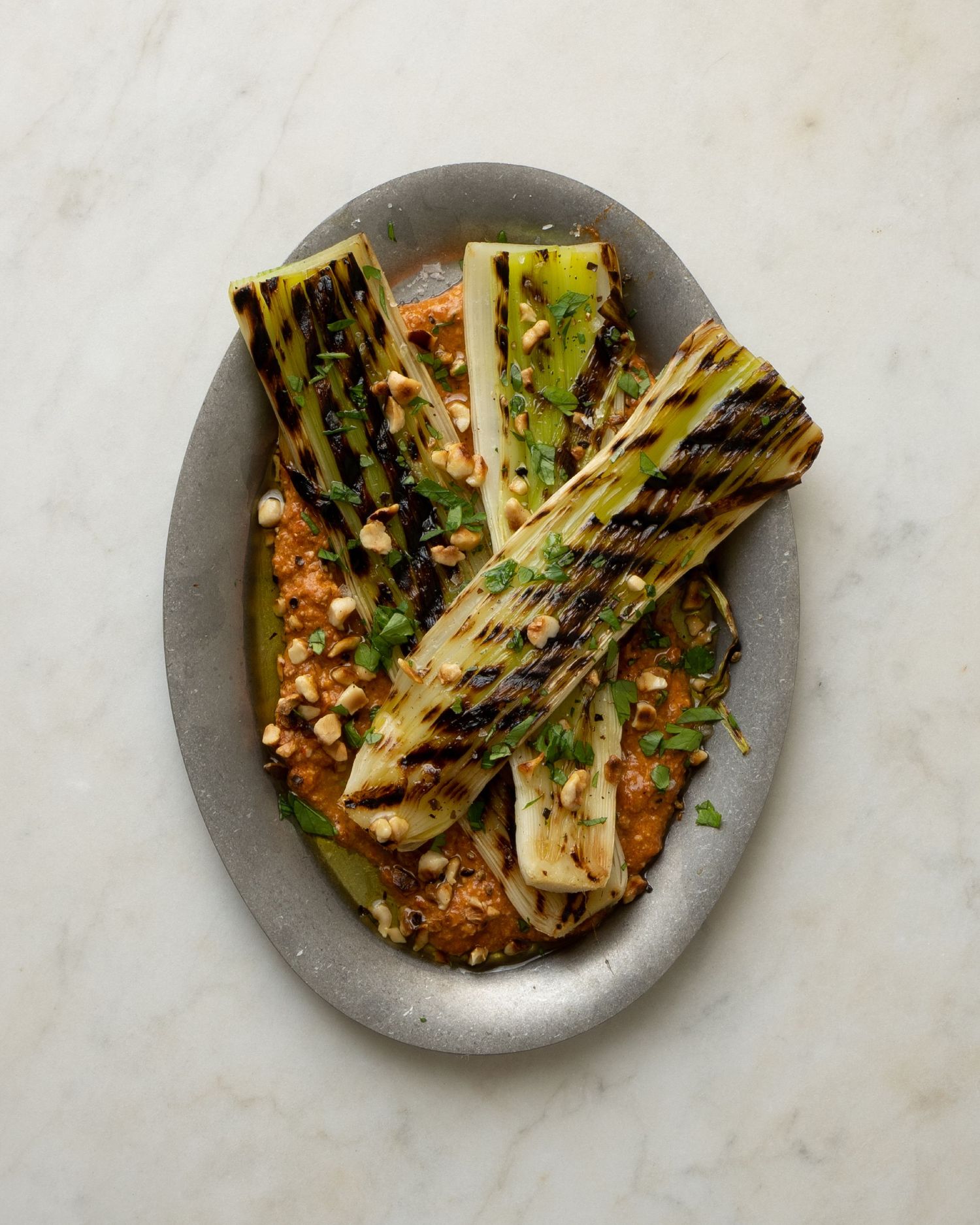 Chargrilled leeks served over romesco sauce with crushed peanuts and herbs on a grey oval plate.