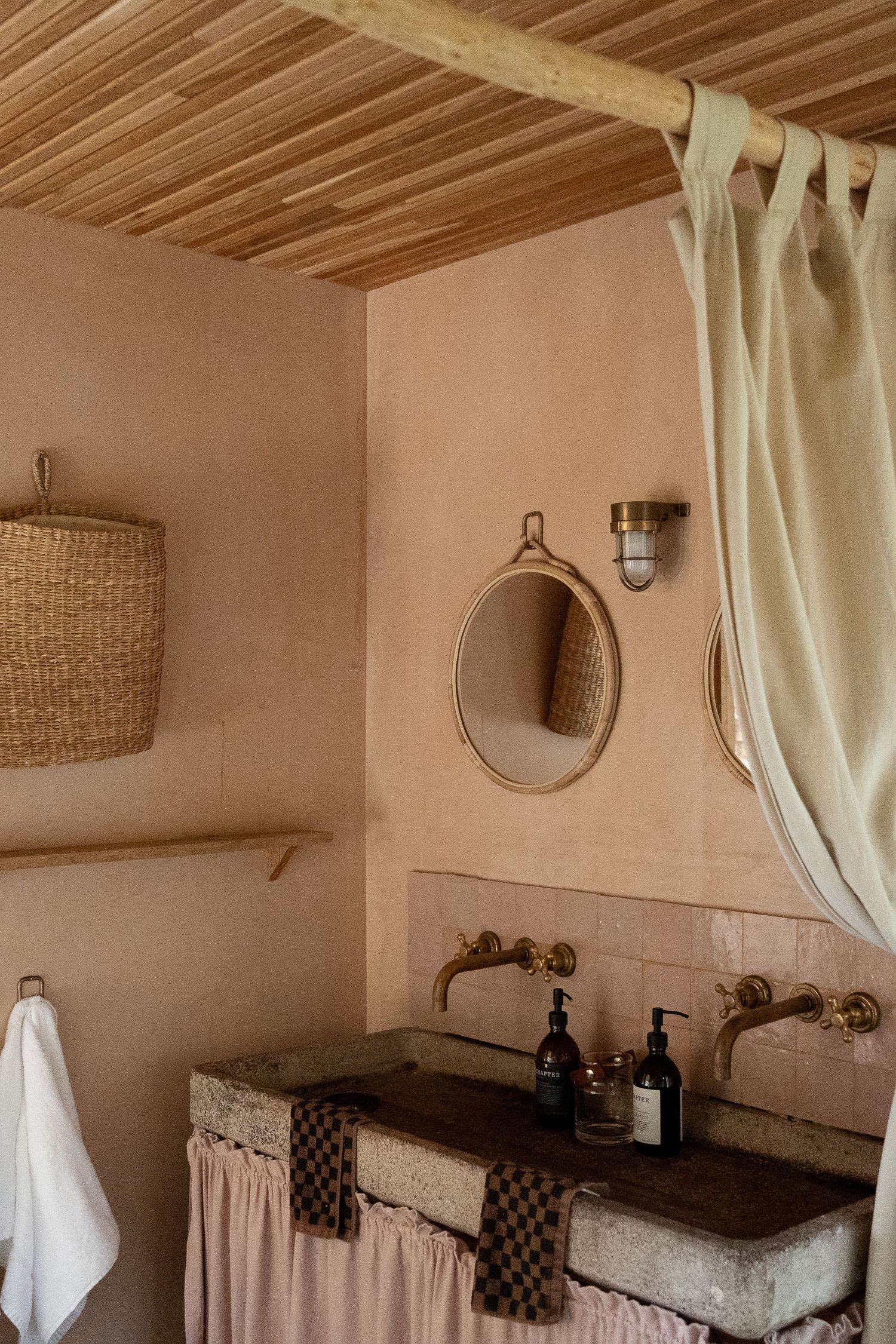Rustic pink plaster bathroom with stone sink, brass fixtures, oval mirror, and wooden slatted ceiling.