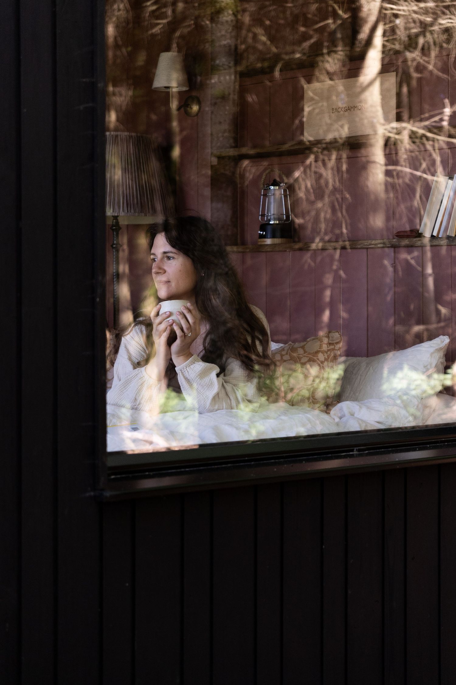 Woman holding a cup smiling while relaxing in bed, photographed through a window reflecting forest trees.