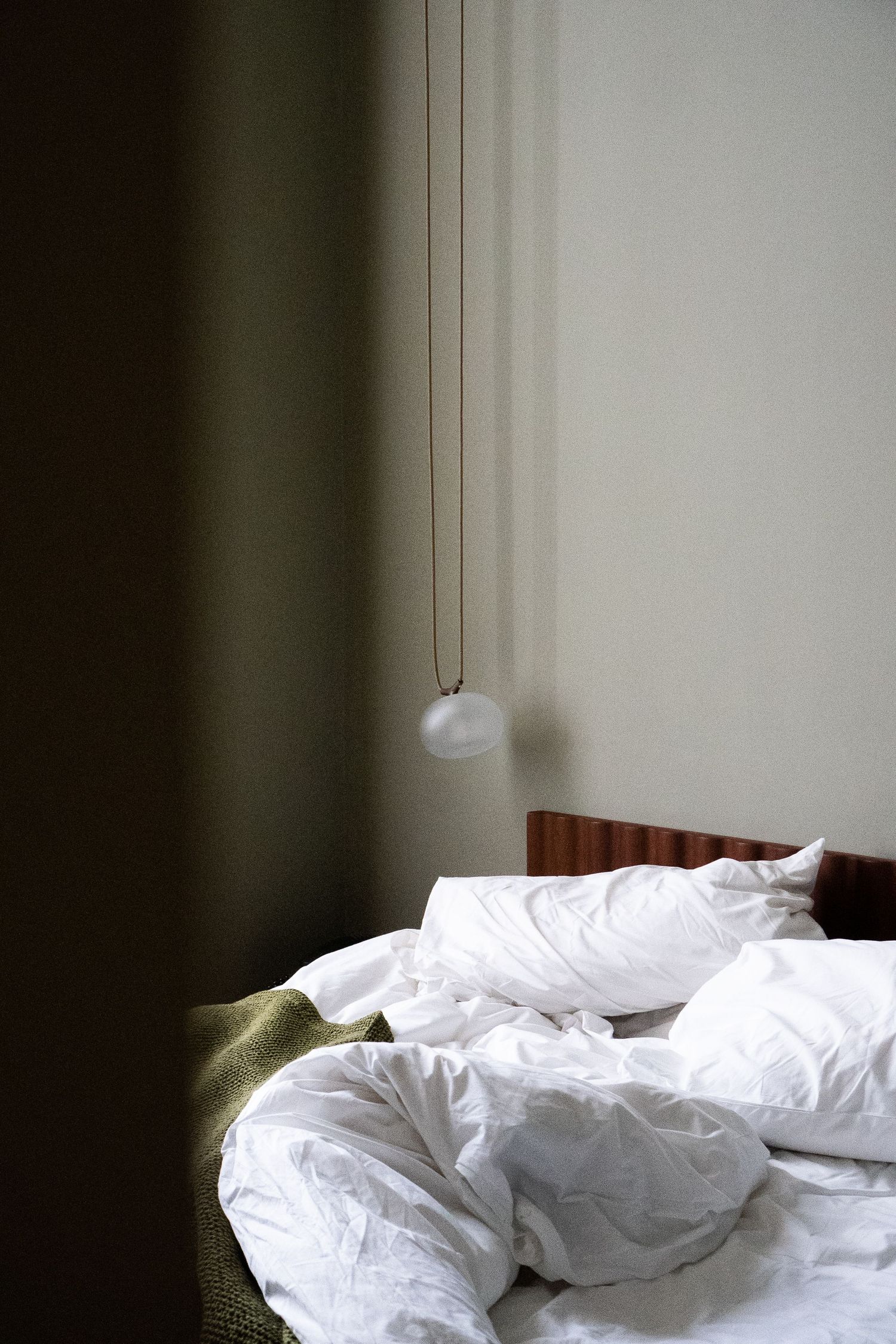 Unmade bed with white linen pillows and duvet, wooden headboard, and delicate pendant light in dim bedroom.