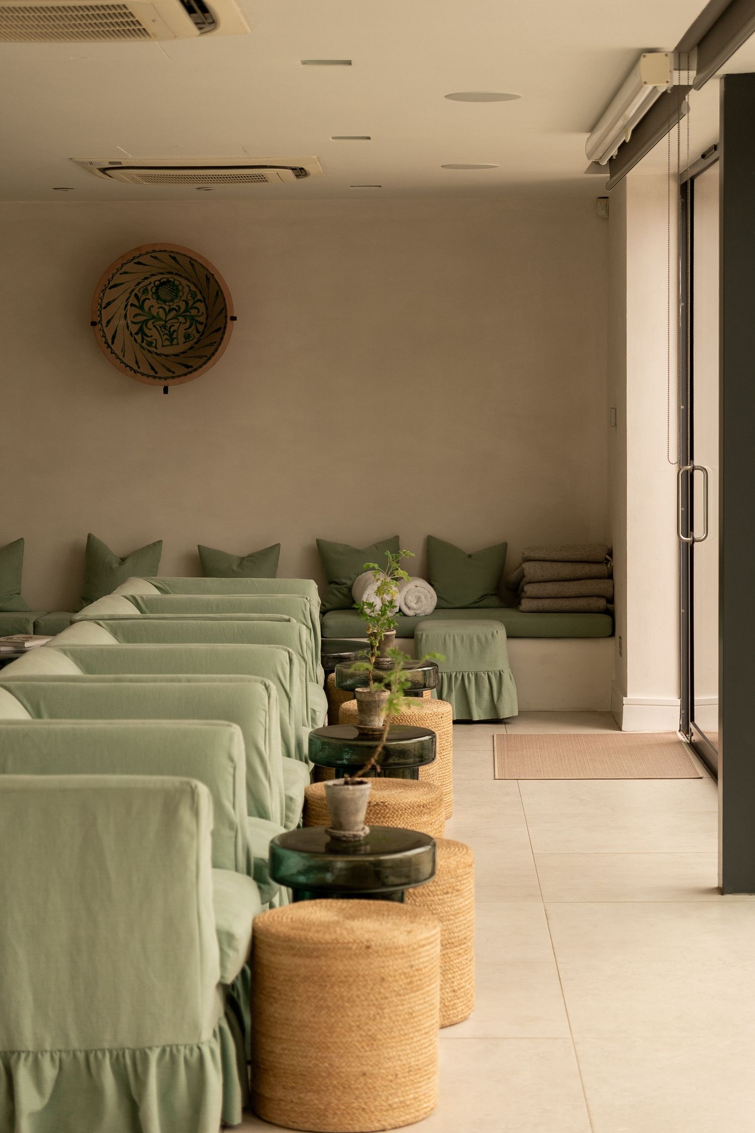 Serene spa relaxation room with sage green lounge chairs, wicker stools, and potted plants in a minimalist setting.