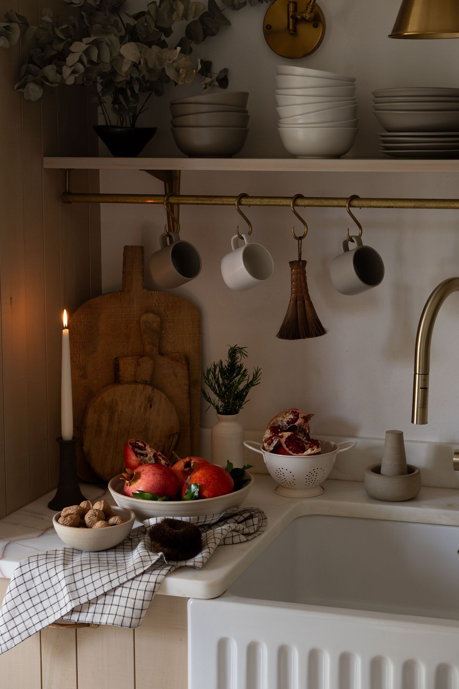 Cozy kitchen with brass fixtures, hanging mugs, pomegranates, candlelight, and rustic wooden cutting boards.