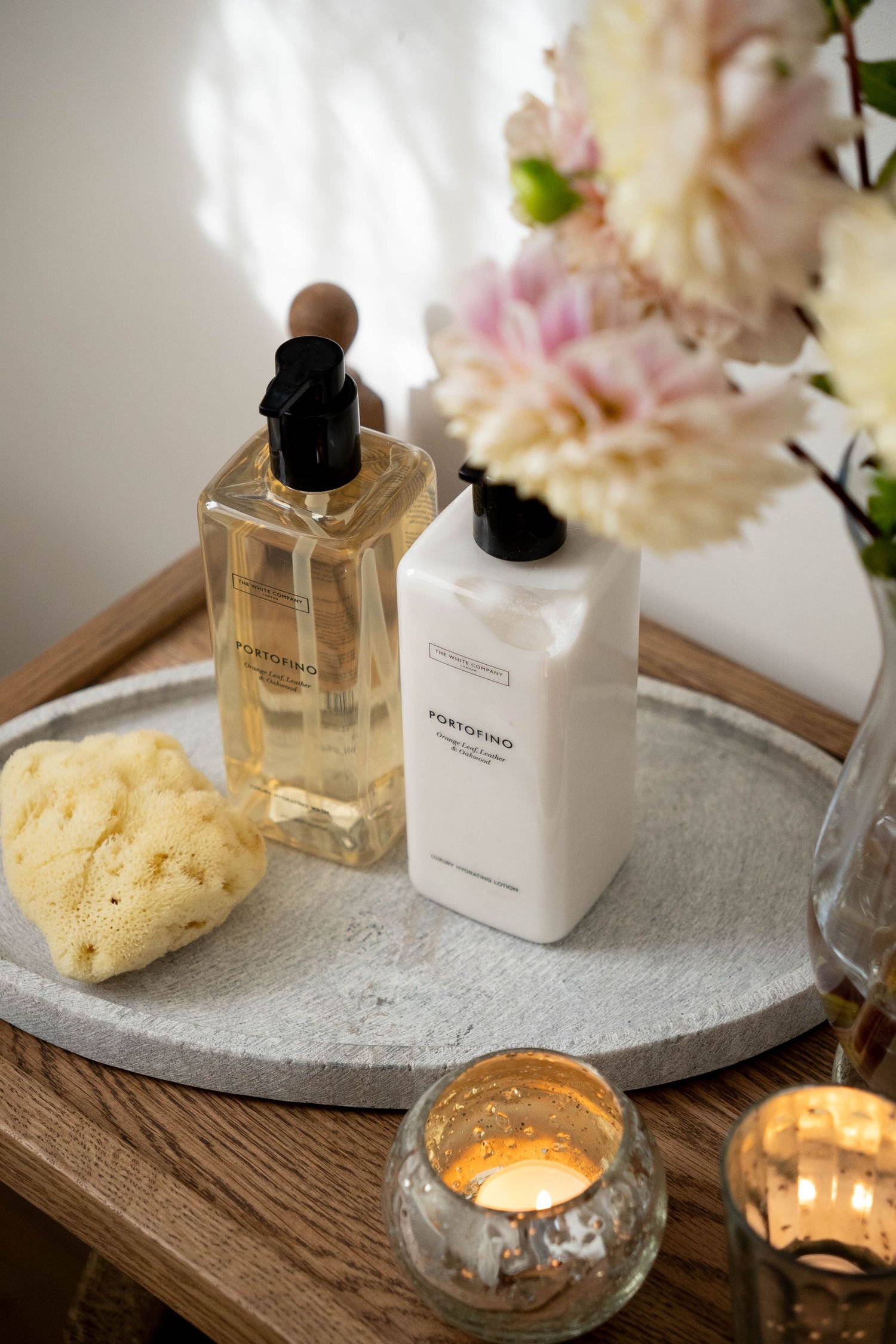 Portofino hand wash and lotion bottles on a stone tray with a sponge, lit candle, and fresh flowers.