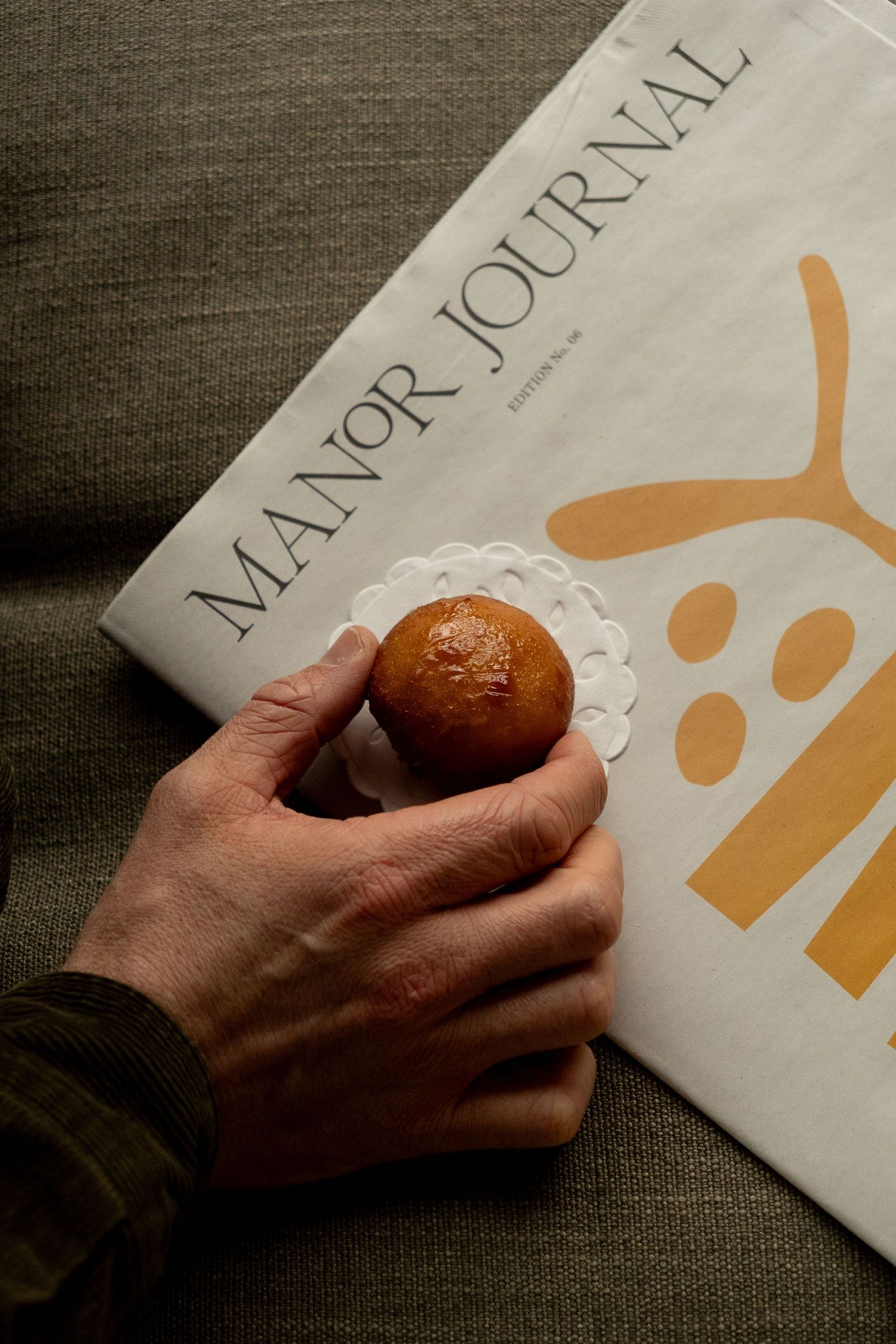 Hand holding a glazed brioche bun on a doily next to a Manor Journal magazine on a linen surface.