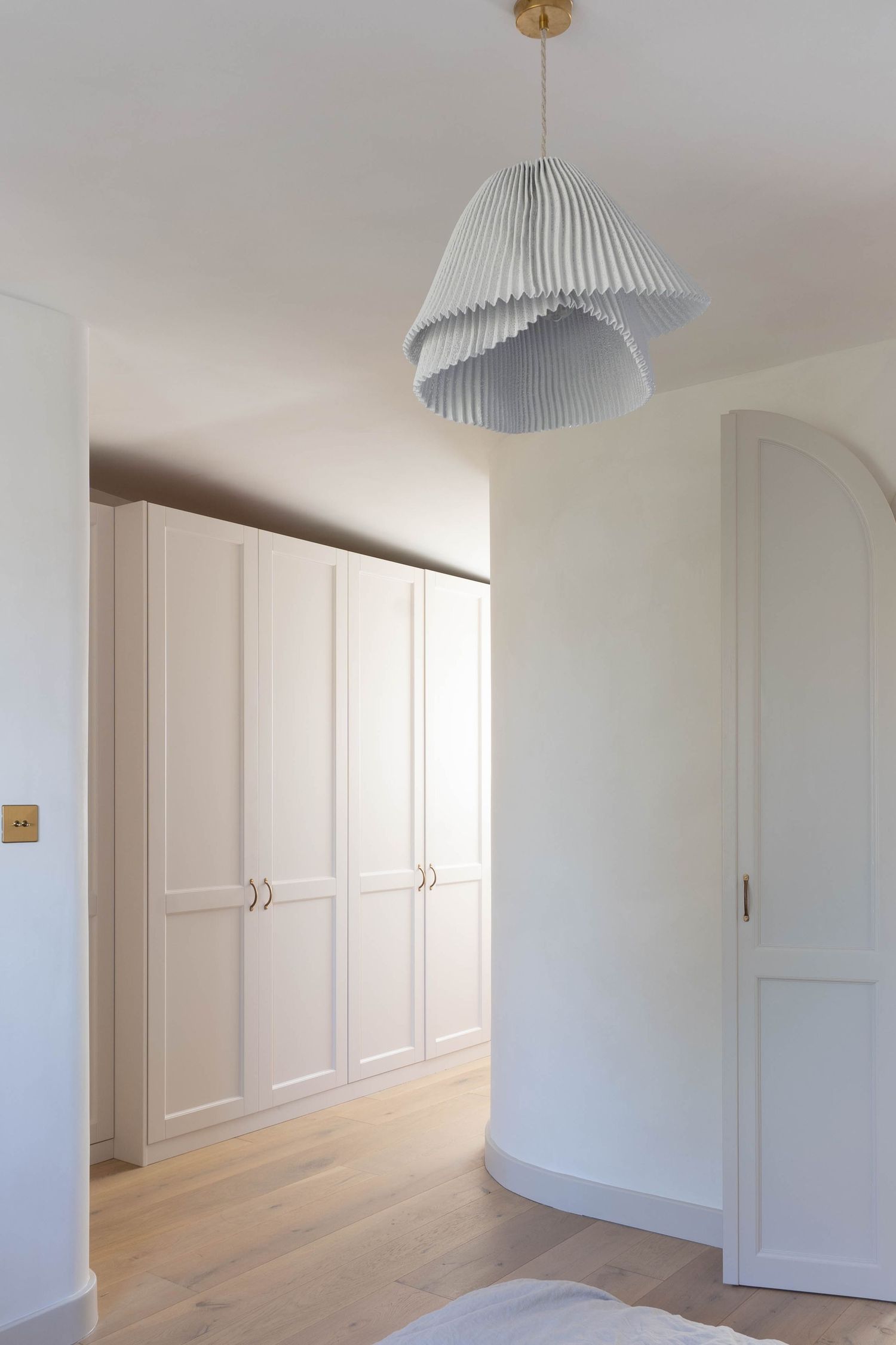 Minimalist bedroom with white built-in wardrobes, light wood floors, and a pleated pendant light in soft neutral tones.