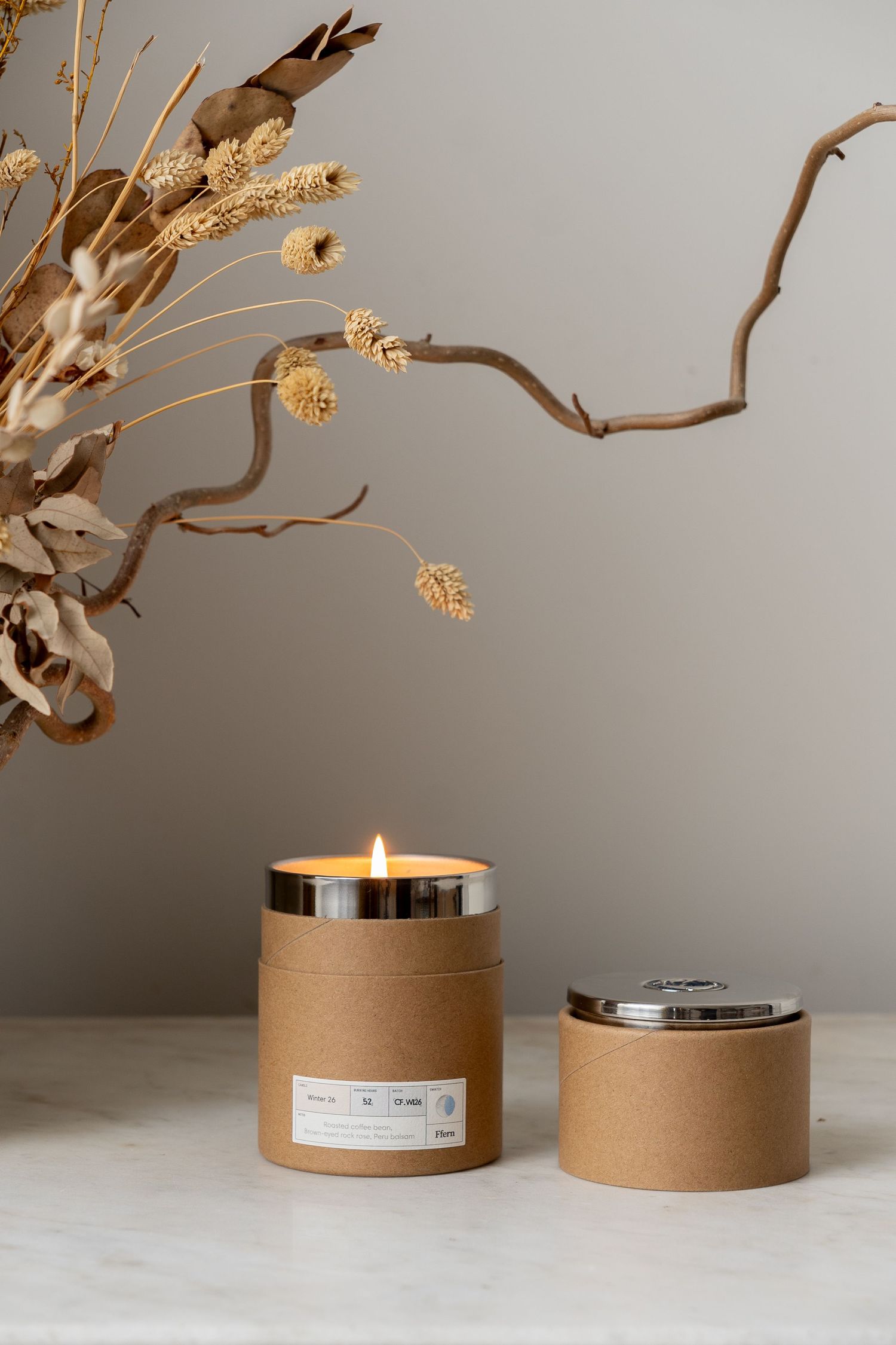 Lit kraft paper candle in silver lid container beside dried botanicals on marble surface.