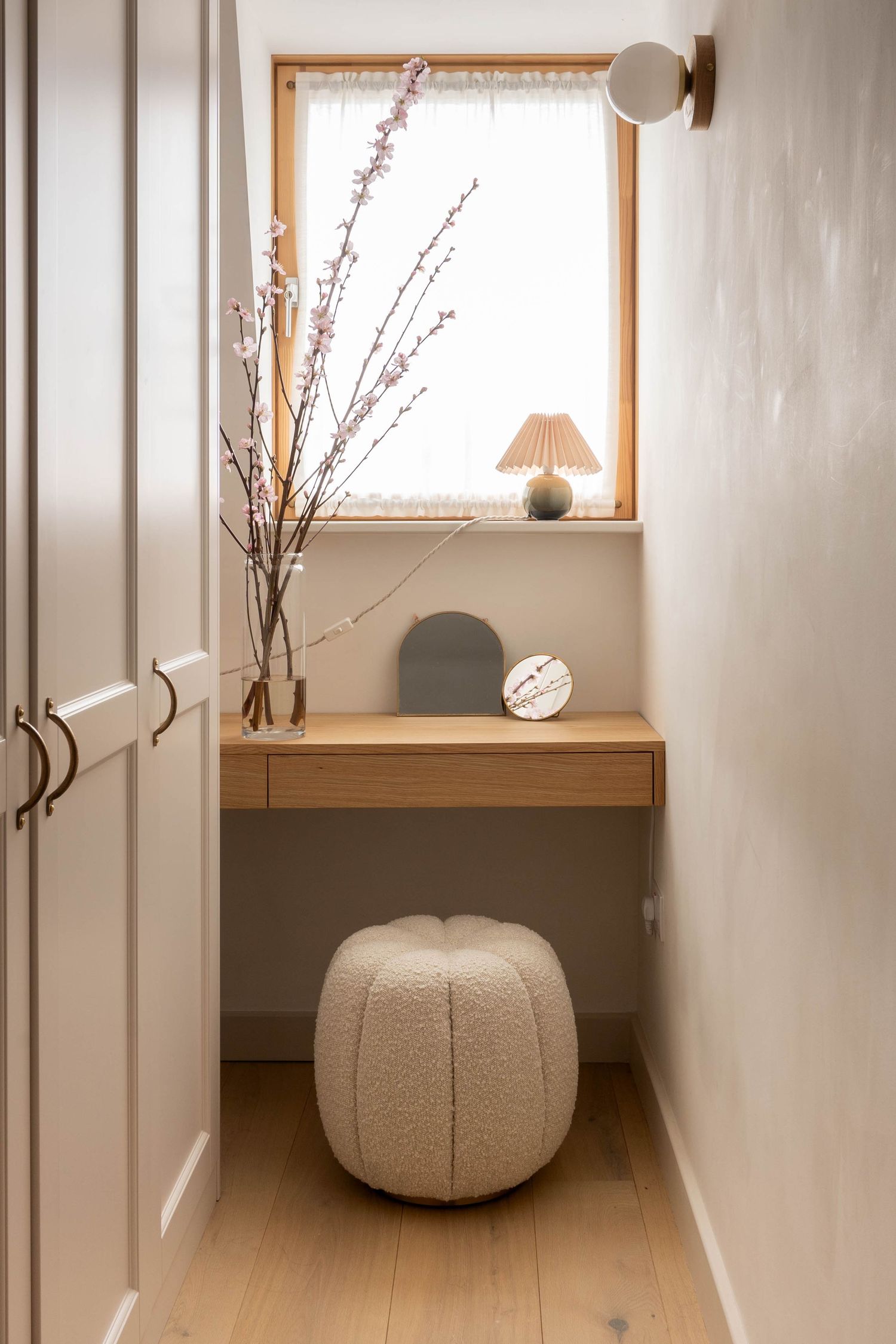 Compact dressing nook with floating oak desk, boucle pouf, cherry blossom branches, and small table lamp by window.