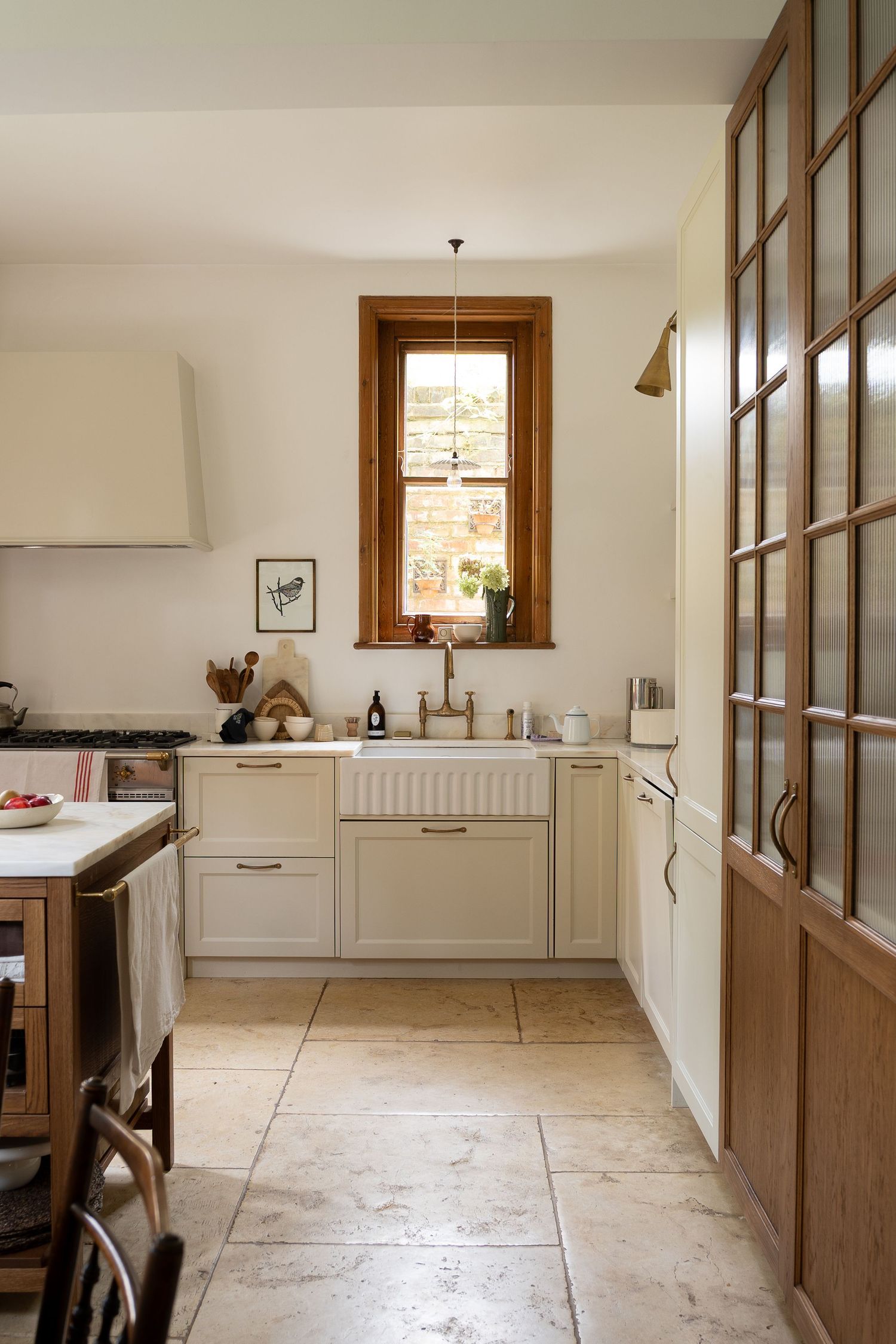 Bright neutral kitchen with cream cabinets, farmhouse sink, wood-framed window, and limestone tile floor.