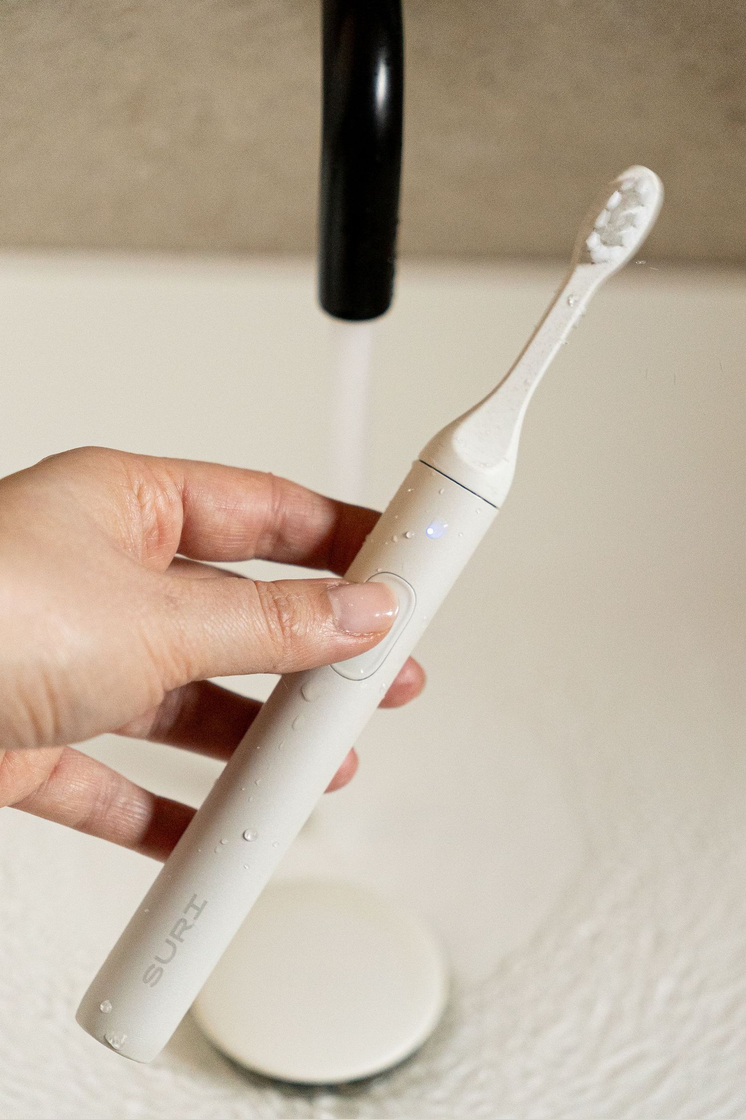 Hand holding a white SURI electric toothbrush under running water at a bathroom sink.