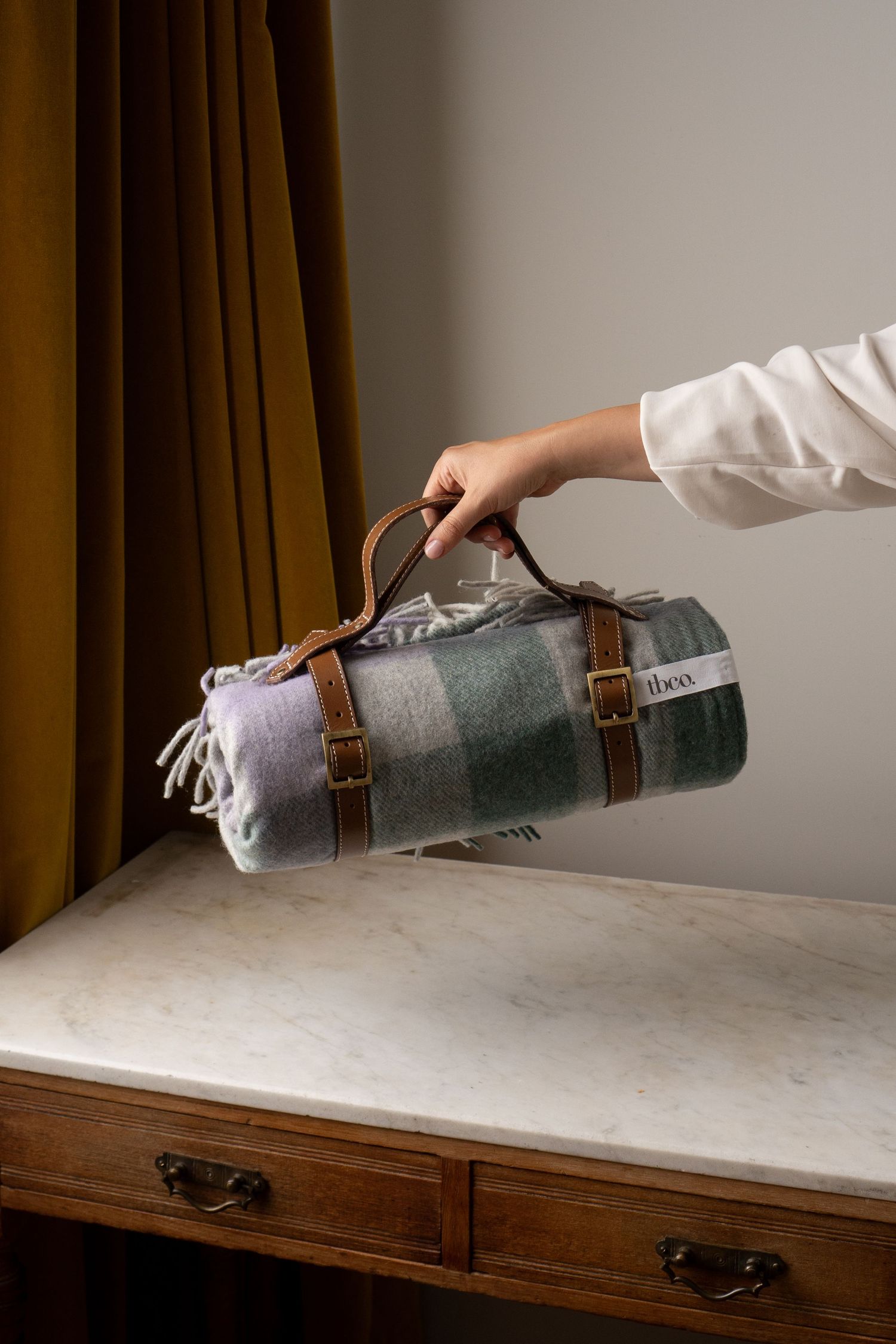 Hand holding a rolled green plaid wool blanket with brown leather carry strap over a marble dresser.