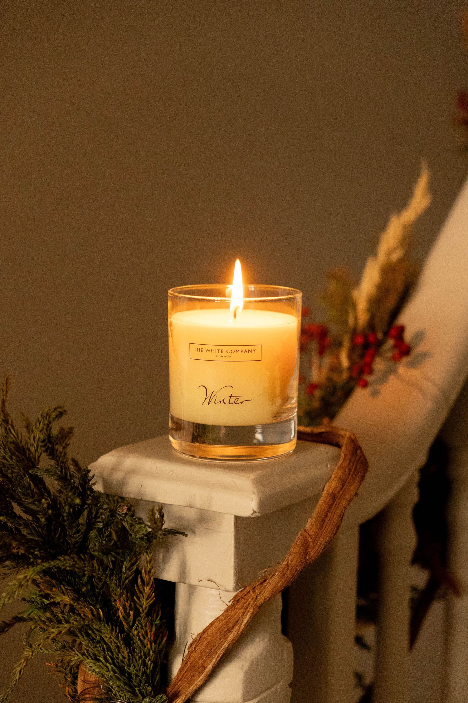 The White Company Winter scented candle glowing warmly on a white stair post with festive greenery.