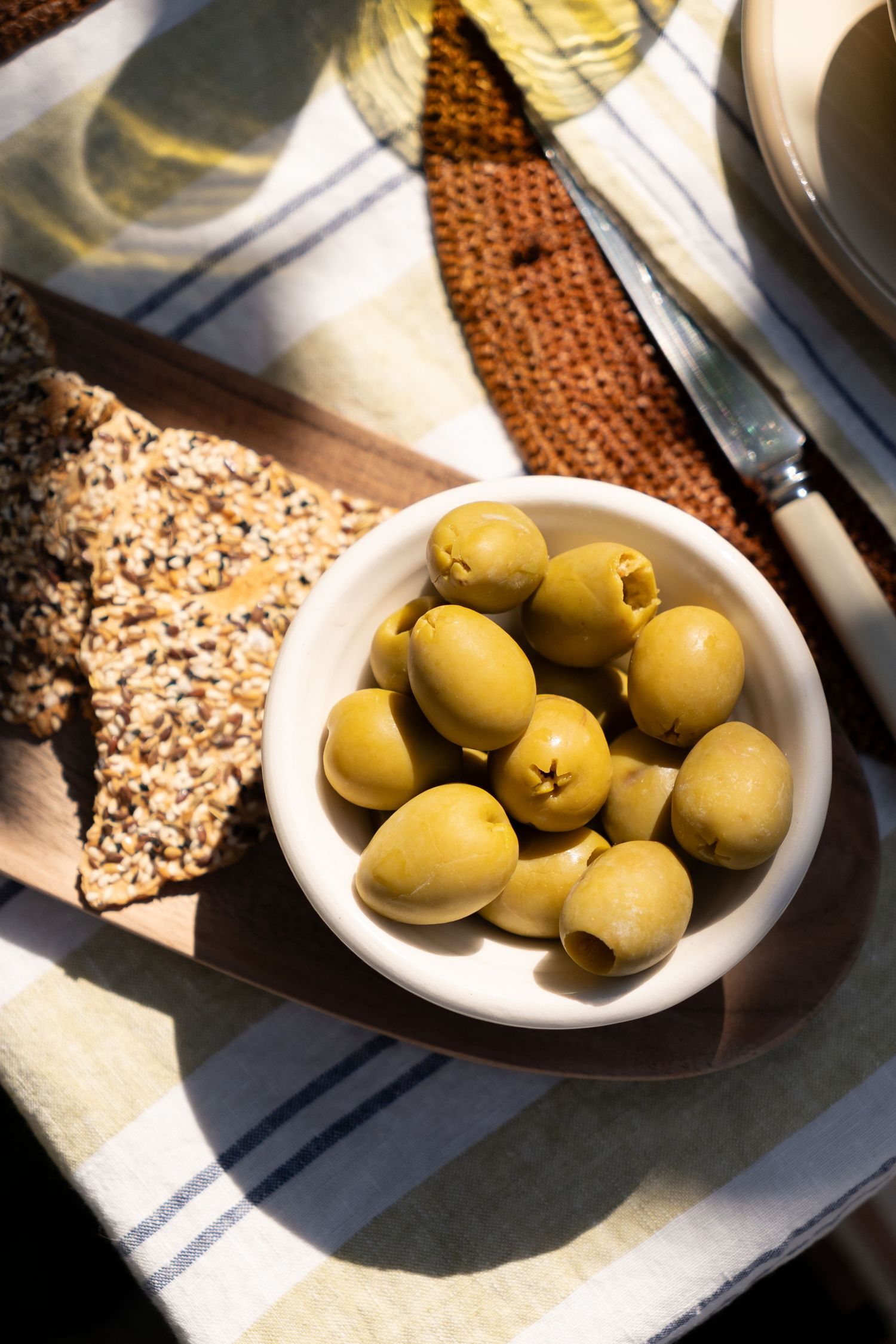 Bowl of green pitted olives served with seeded crackers on a wooden board in bright sunlight.