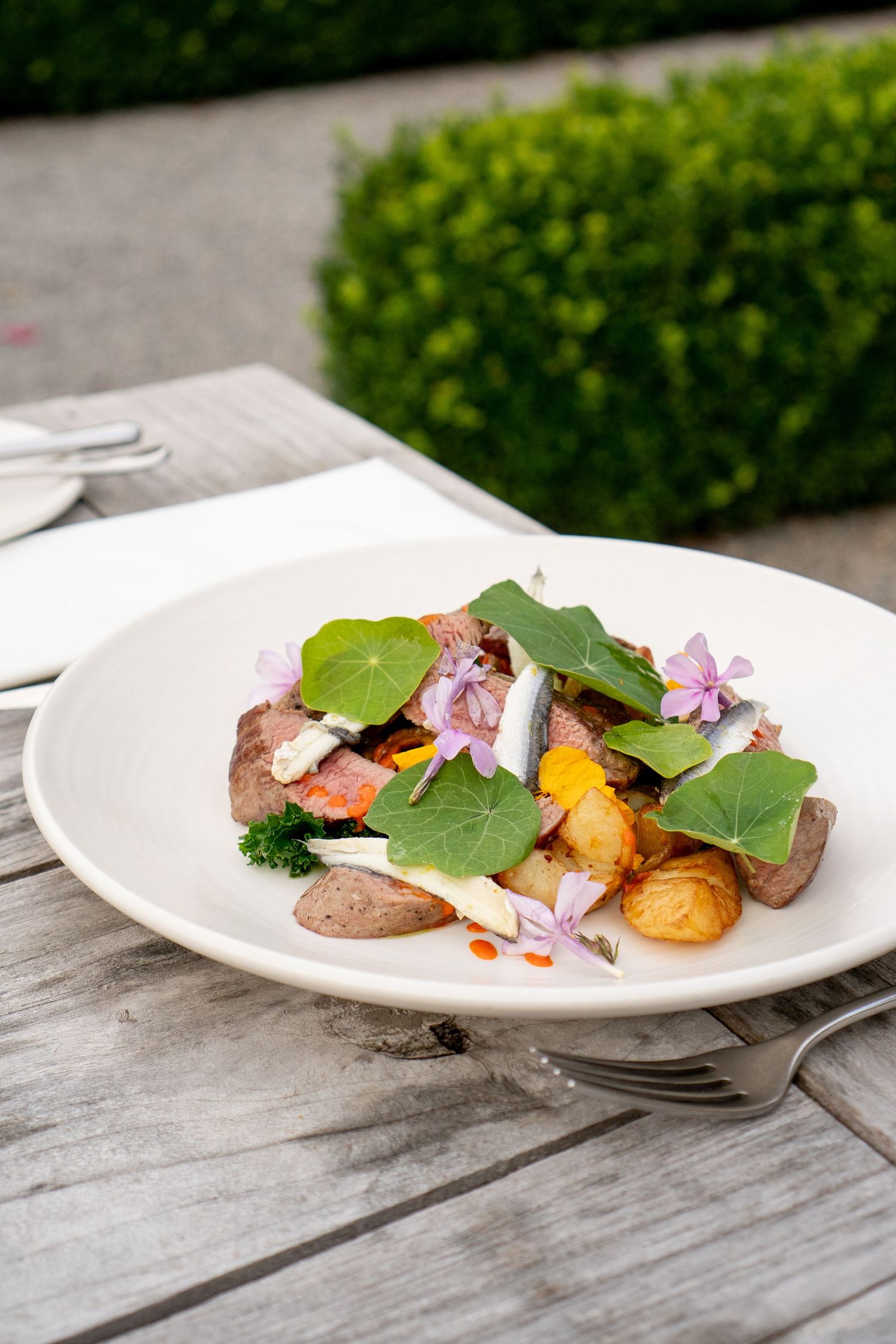 A gourmet dish of roasted potatoes and meat topped with edible flowers and fresh greens served on a white plate outdoors.