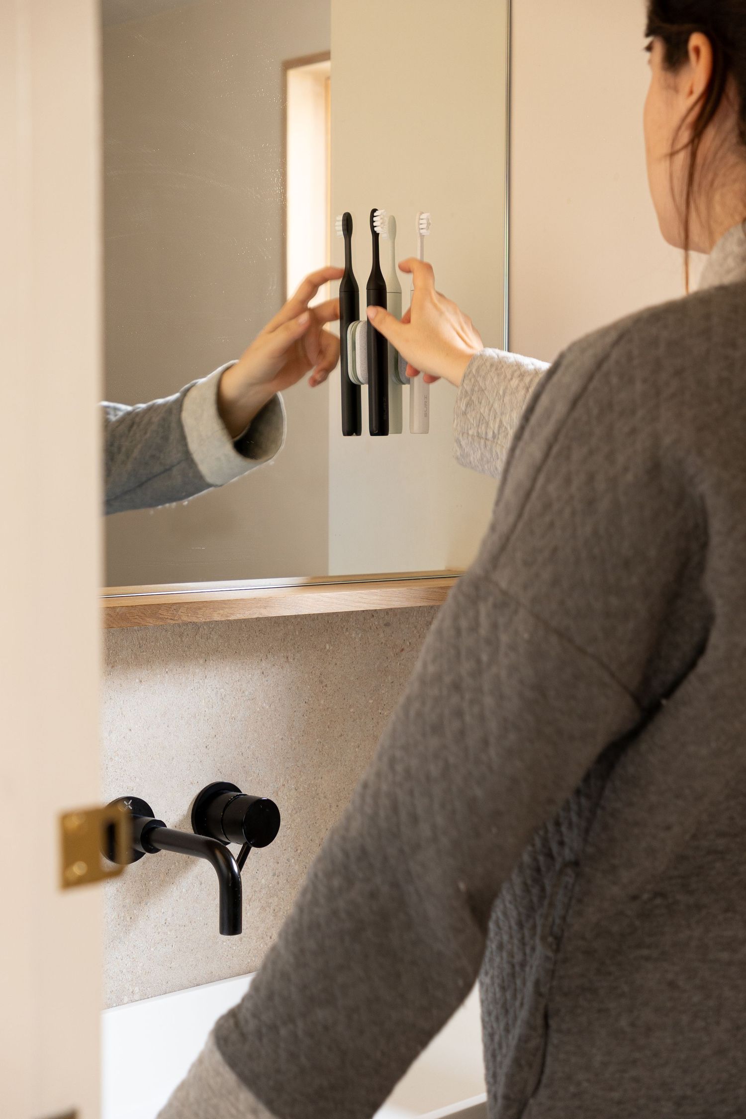 Woman in grey sweater placing a black electric toothbrush on a mirror-mounted holder.