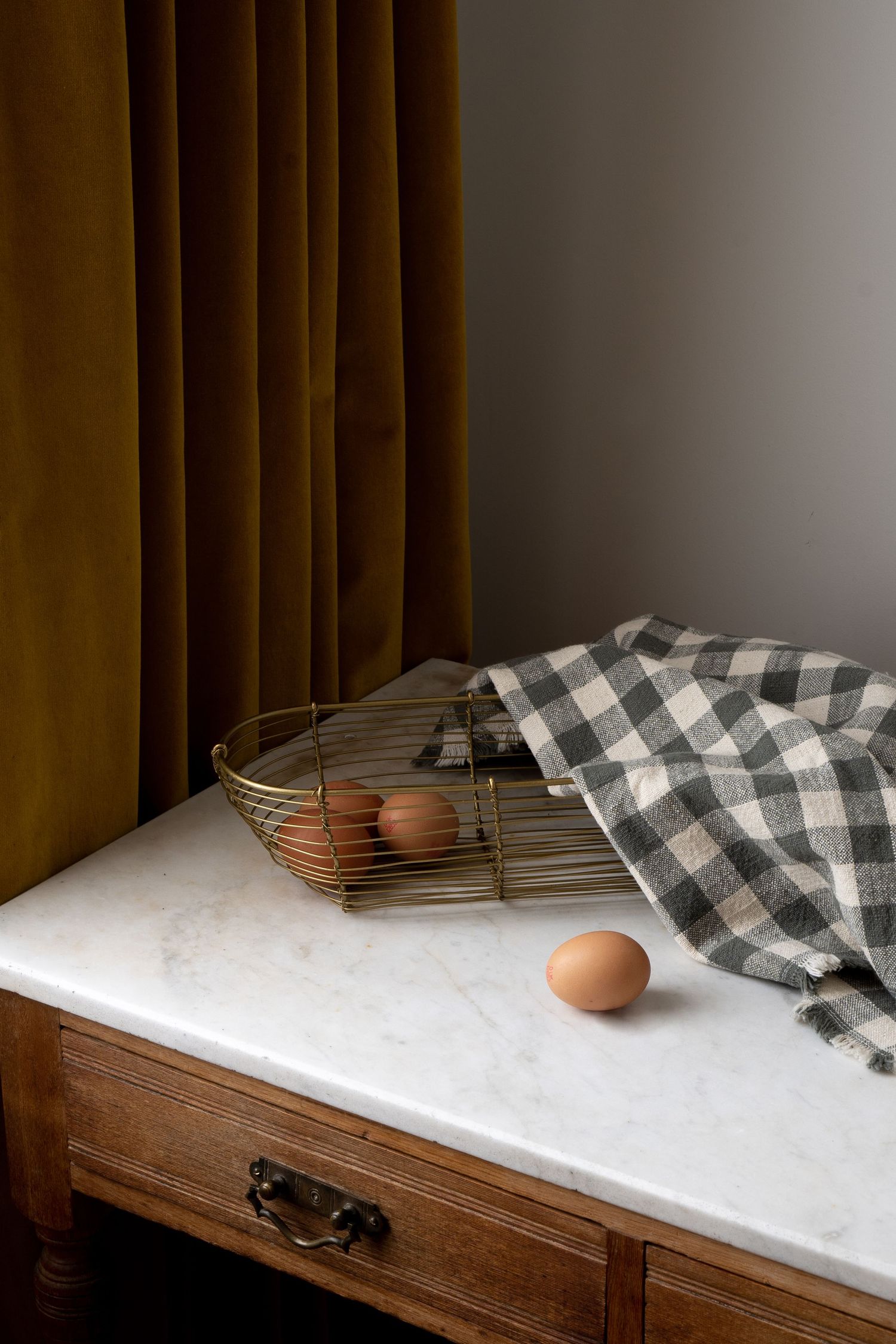 Brown eggs in a wire basket with checkered cloth on a marble-topped wooden dresser.