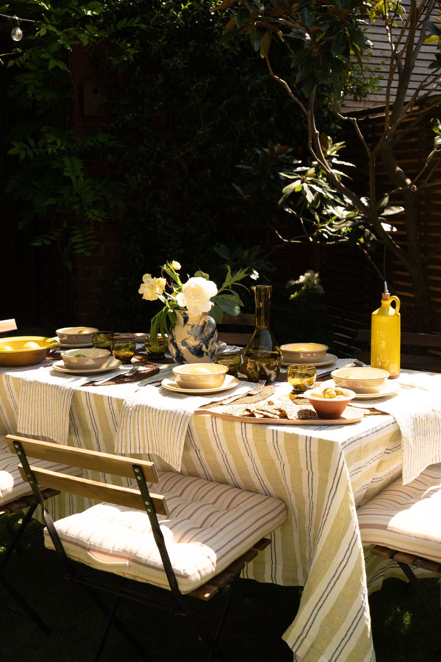 Outdoor dining table set with striped linen tablecloth, ceramic dishes, yellow vase, and white flowers in a sunlit garden.