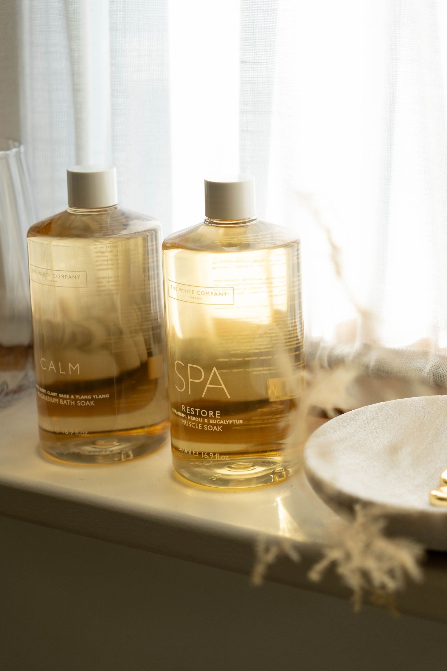 Two spa bath soak bottles labeled Calm and SPA Restore sit on a white shelf in soft natural light.