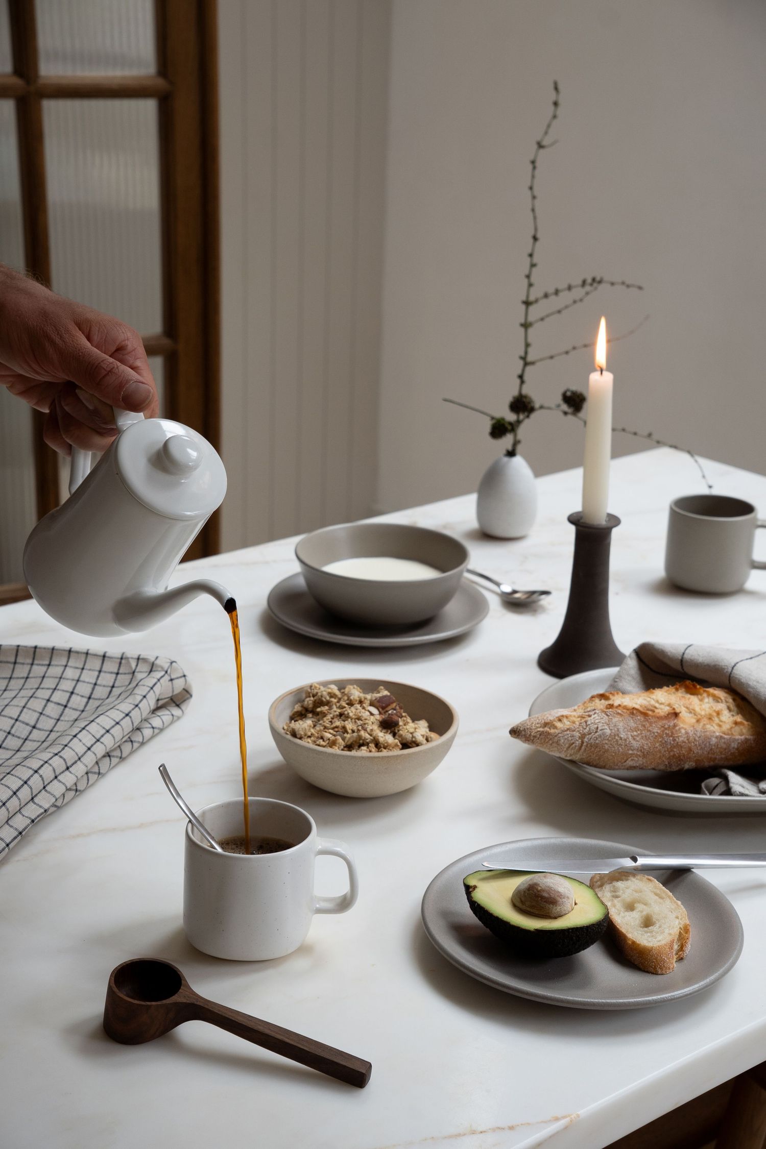 A hand pouring coffee into a mug on a marble table set with avocado toast, oatmeal, bread, and a lit candle.