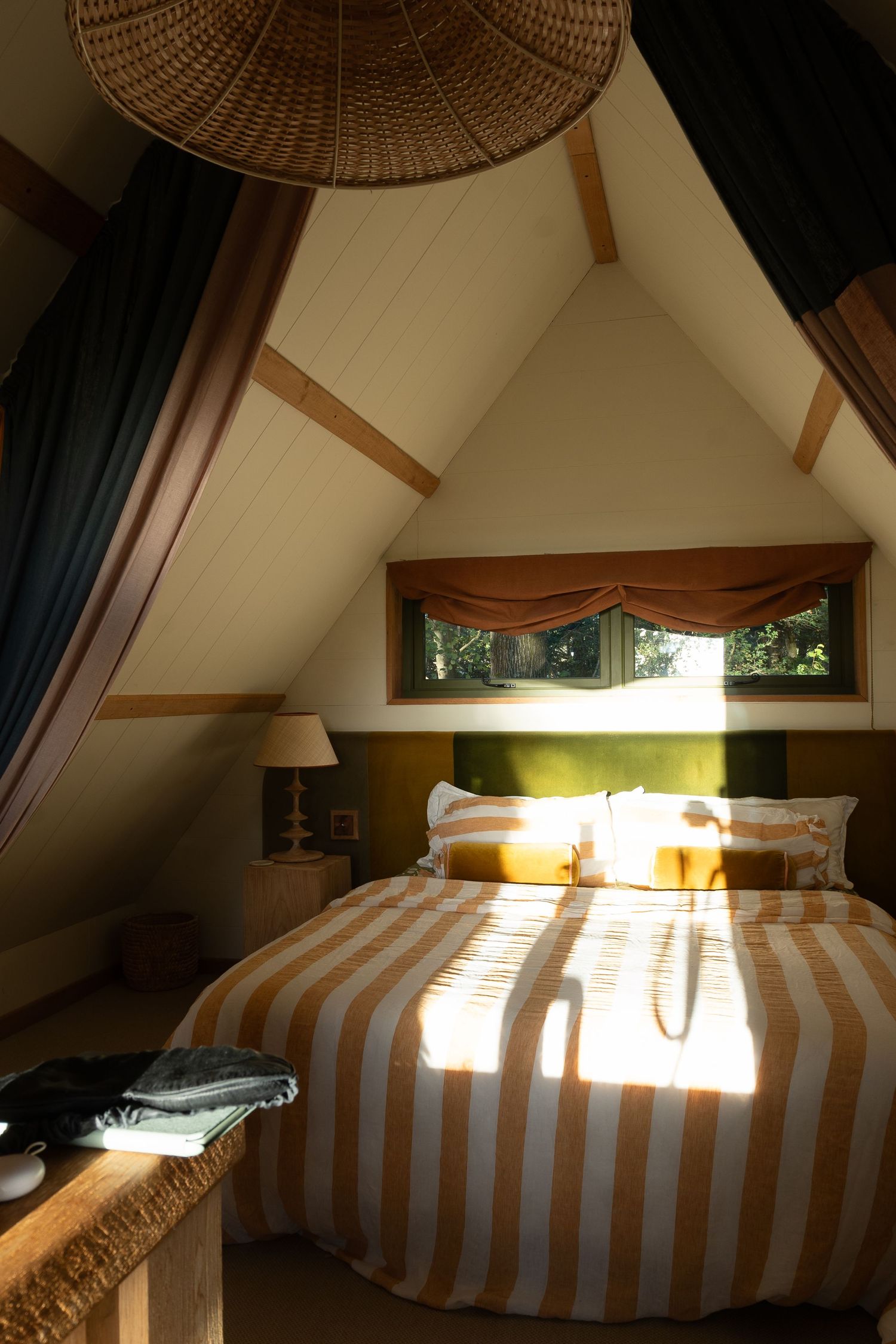 Cozy attic bedroom with striped bedding, triangular wooden beams, and warm sunlight through a small window.