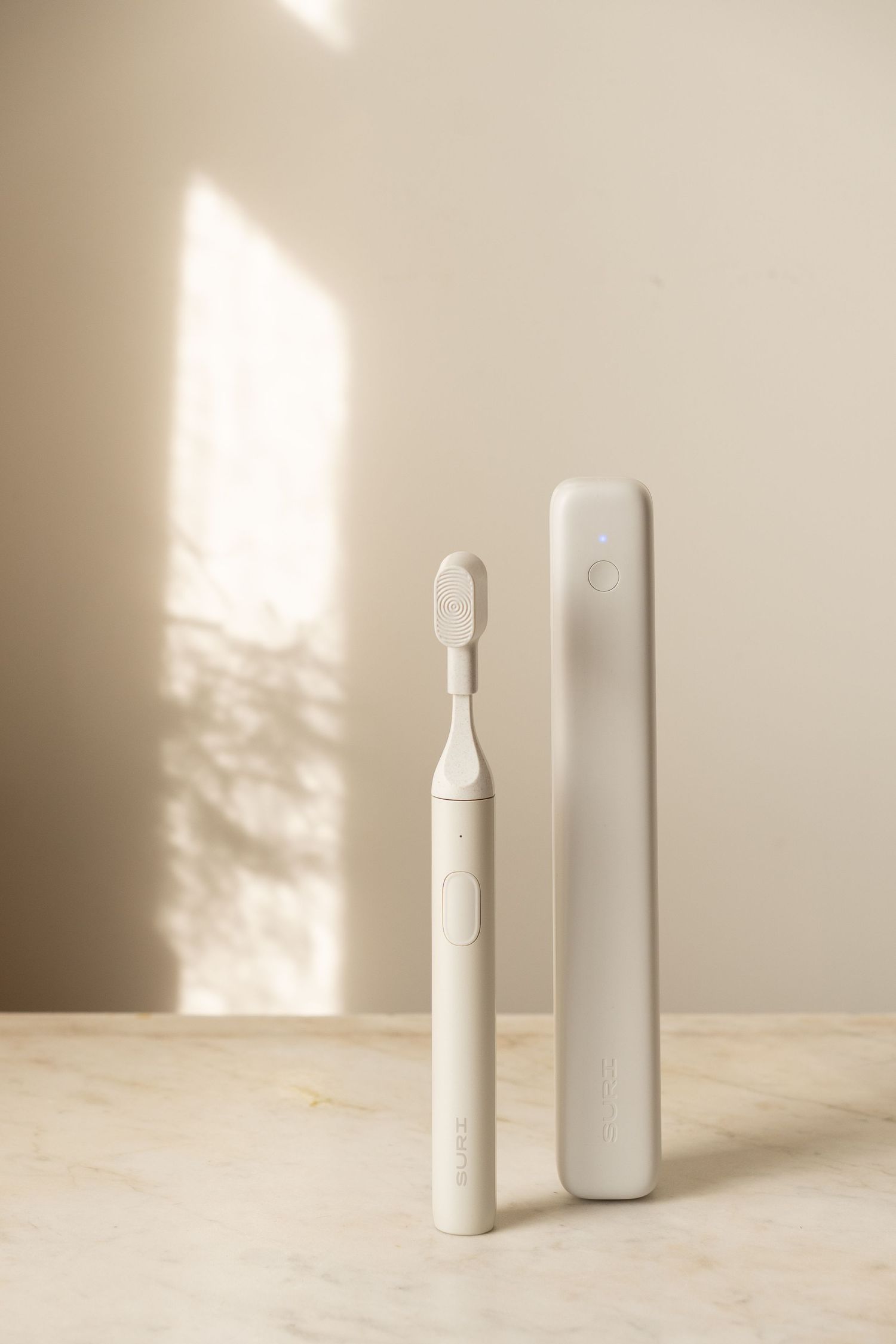 Minimalist white electric toothbrush and travel case on a marble surface with soft shadow light.