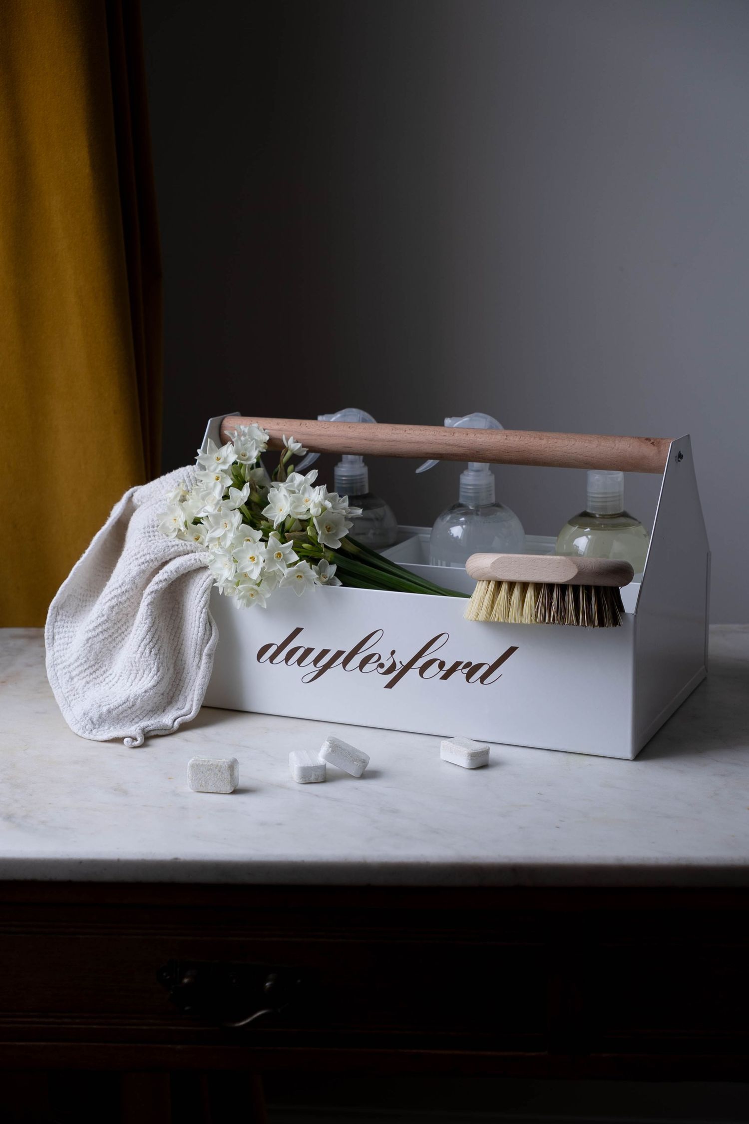 Daylesford white wooden caddy with natural brush, cleaning products, white flowers, and cloth on marble surface.