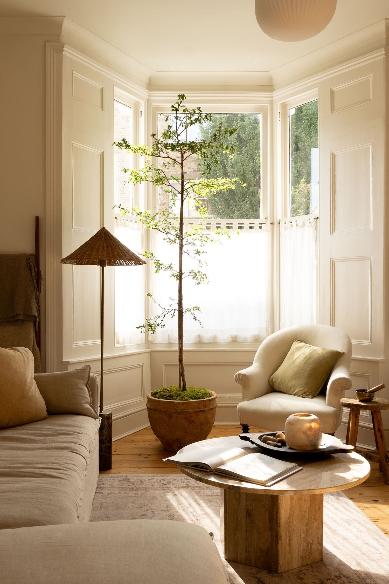 Bright Scandinavian living room with bay window, tall potted tree, cream armchair, round wood coffee table, and floor lamp.