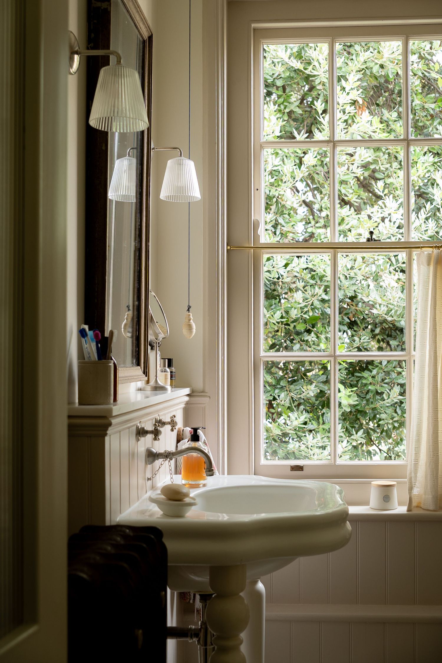 Traditional white pedestal sink with wall-mounted lamp and mirror beside large window with garden views.