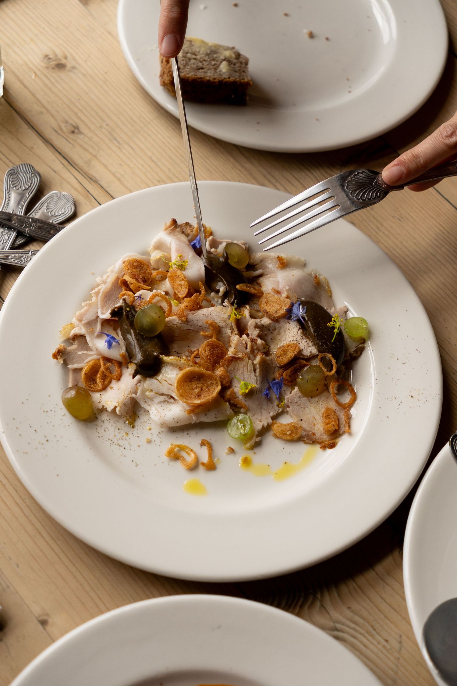 Hand using fork to eat gourmet white fish carpaccio topped with crispy onions, grapes, and edible flowers.