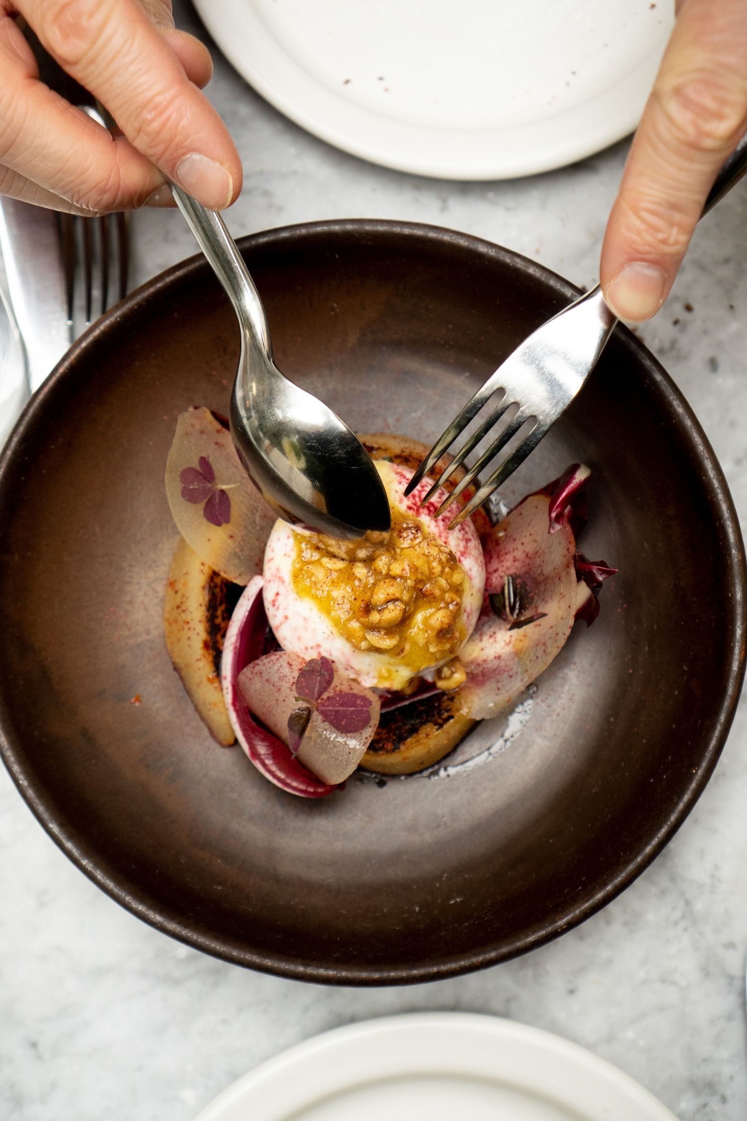 Hands using cutlery to eat elegant radicchio salad with mustard dressing in dark ceramic bowl.