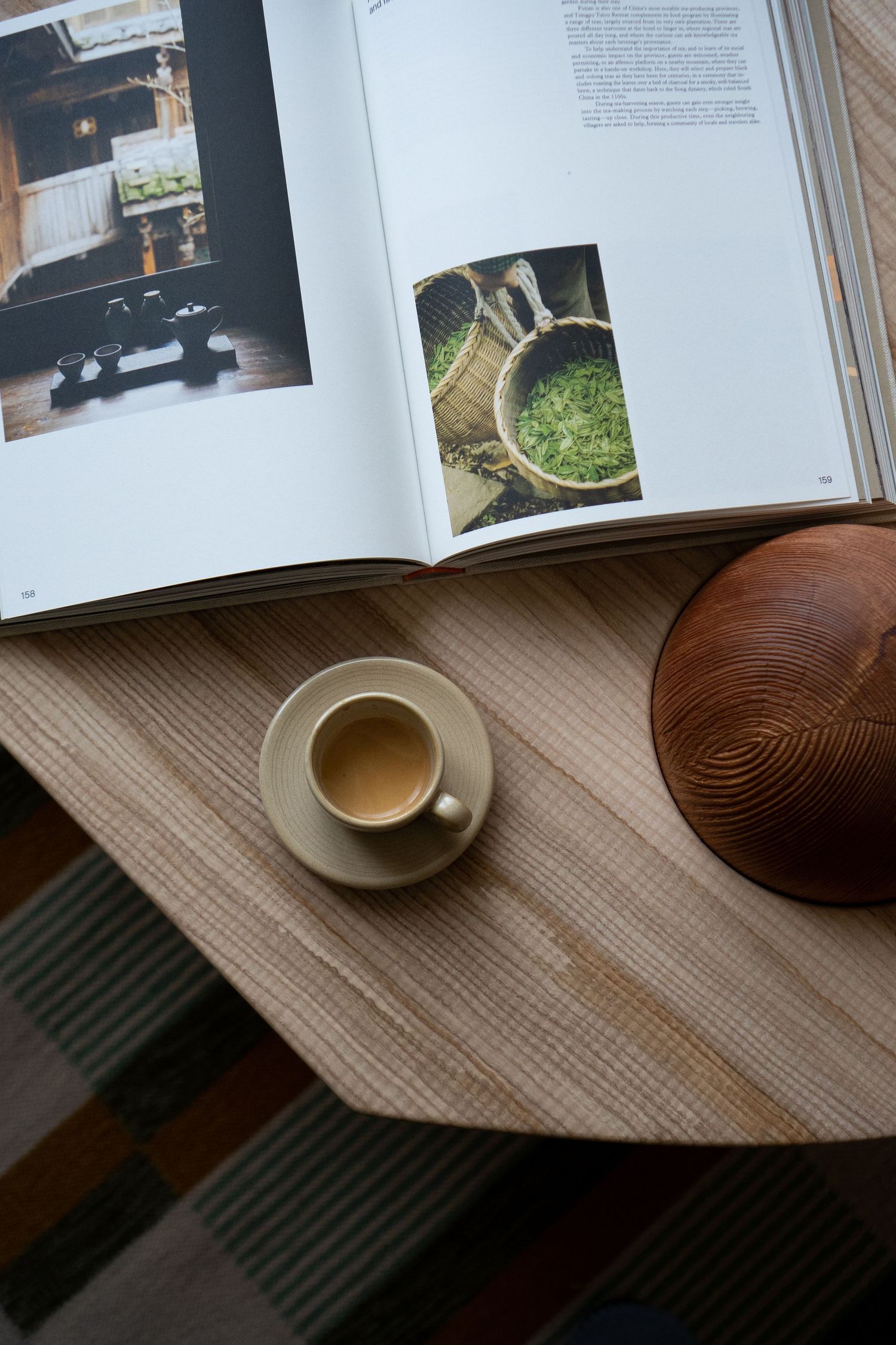 Overhead view of an espresso cup on a saucer beside an open cookbook and wooden bowl on a table.