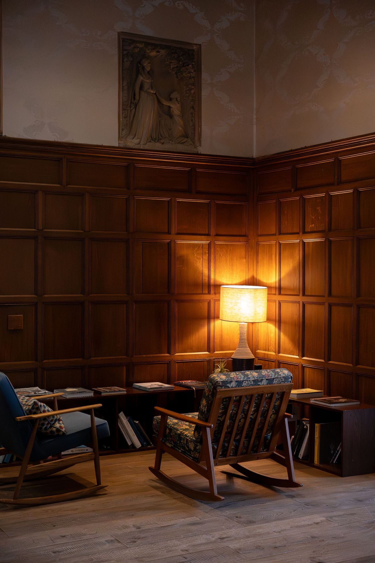 Cozy wood-paneled reading nook with rocking chairs, a glowing table lamp, and low bookshelves on hardwood floors.