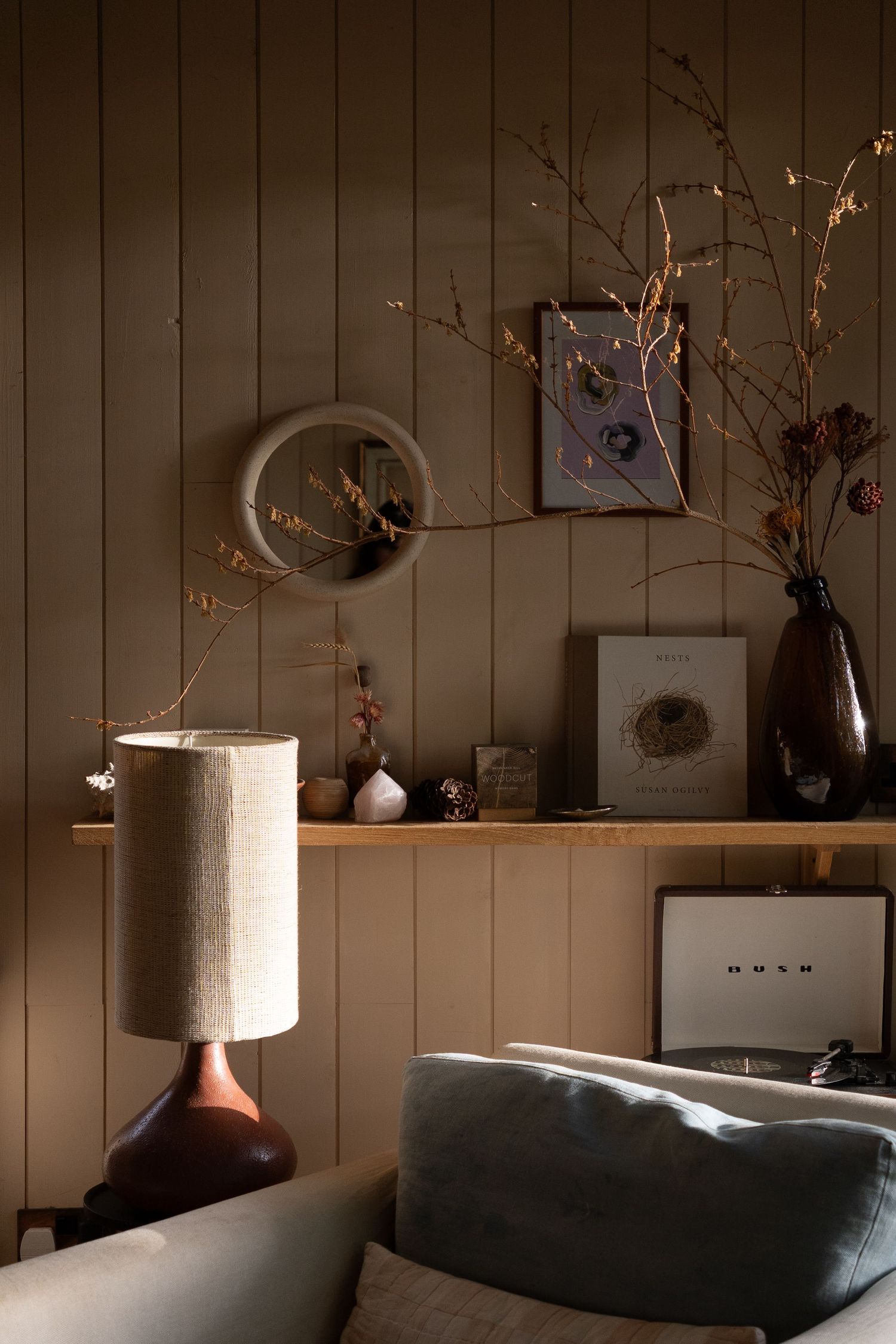 Warm moody living room corner with a ceramic table lamp, dried branches, framed art, and a vinyl record player.