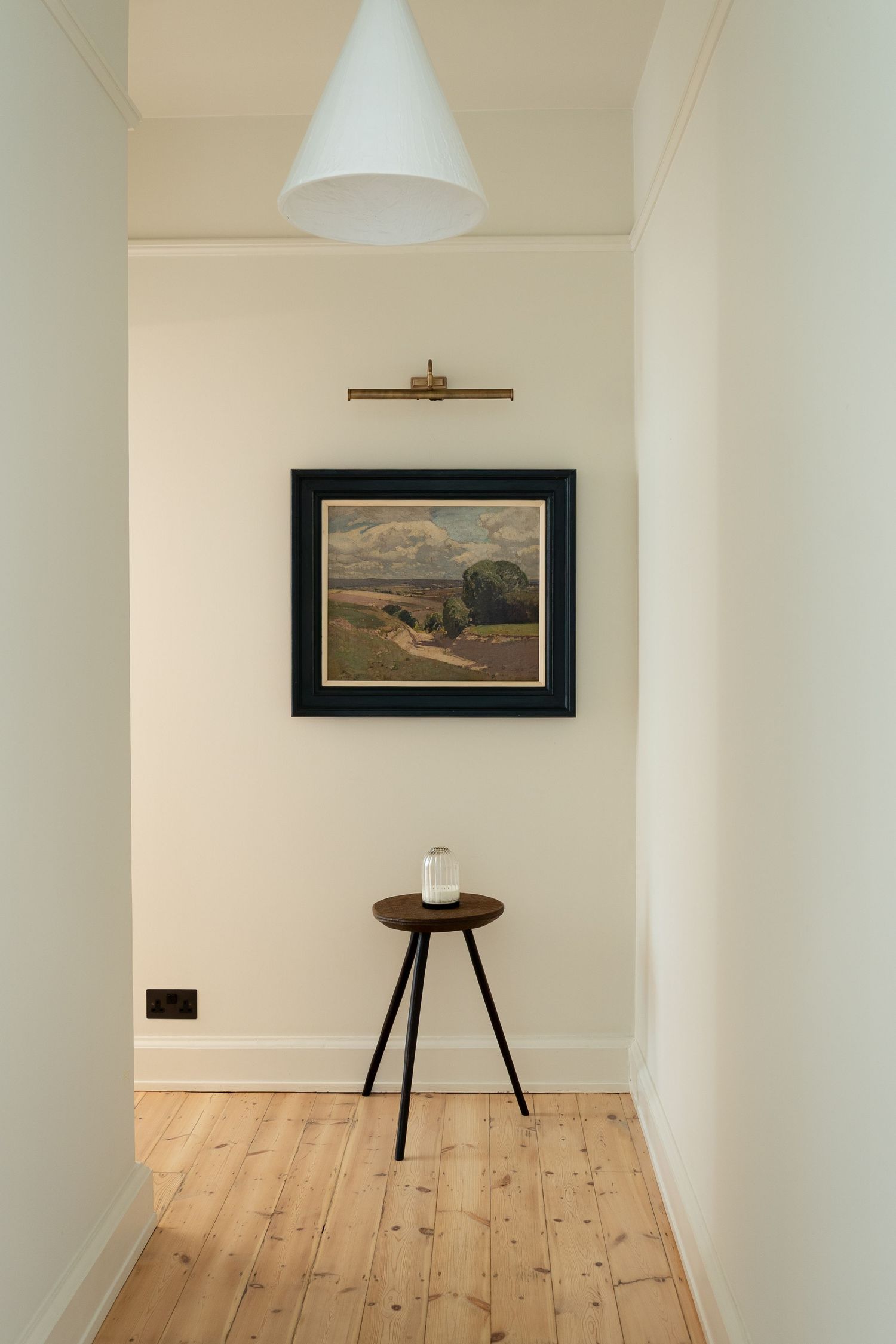 Minimalist hallway with landscape painting, brass picture light, small wooden stool and cone pendant lamp.