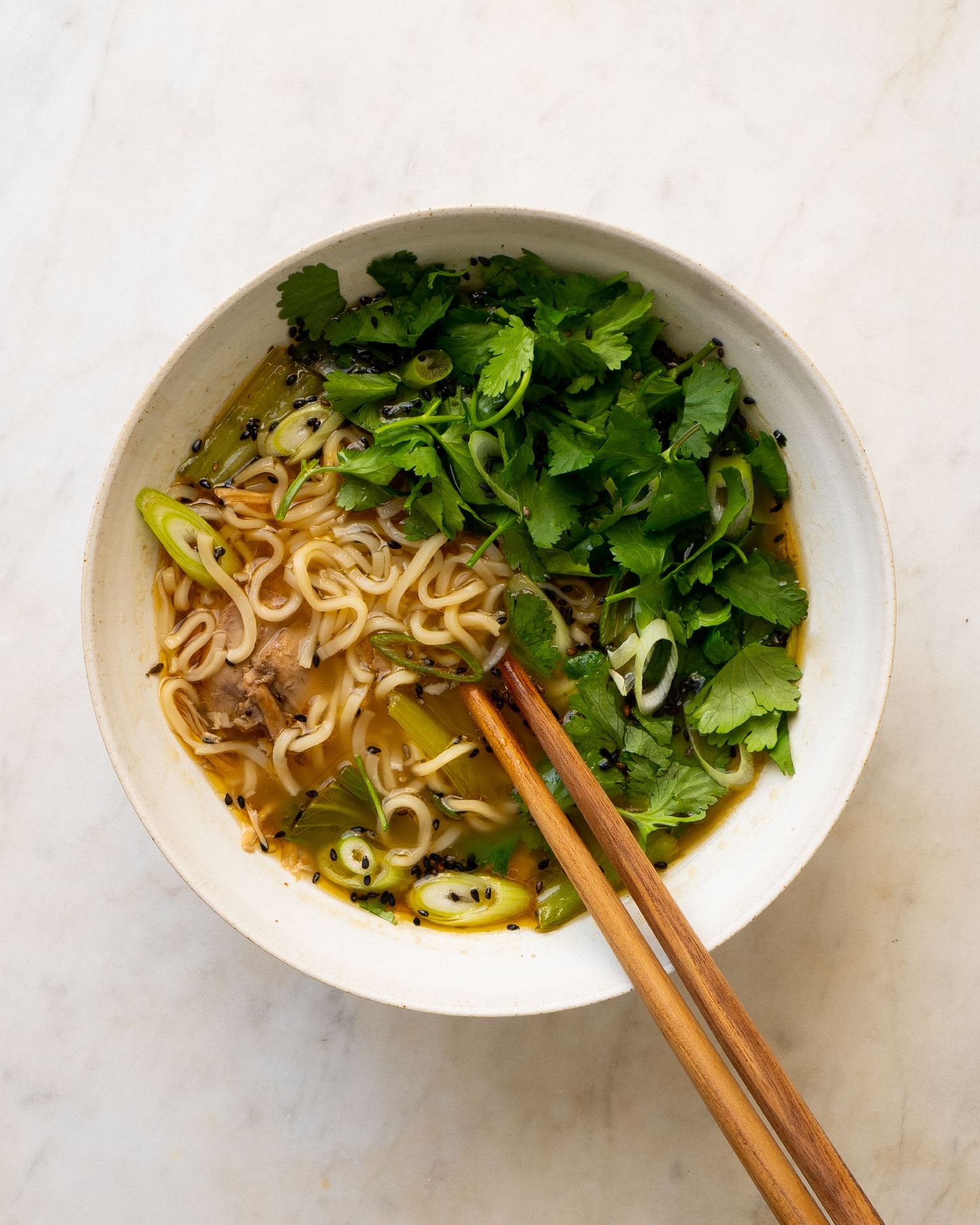 Bowl of ramen noodles in golden broth with fresh cilantro, sliced scallions, and wooden chopsticks.