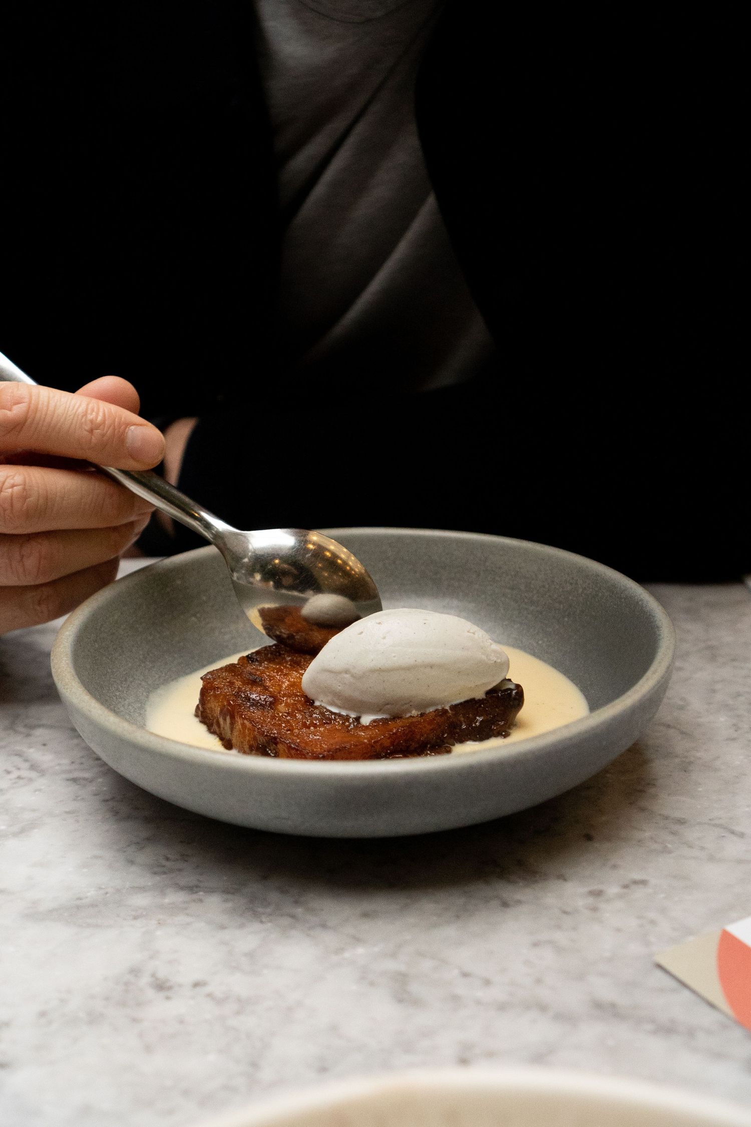Person eating sticky toffee pudding with vanilla ice cream in gray bowl on marble restaurant table.