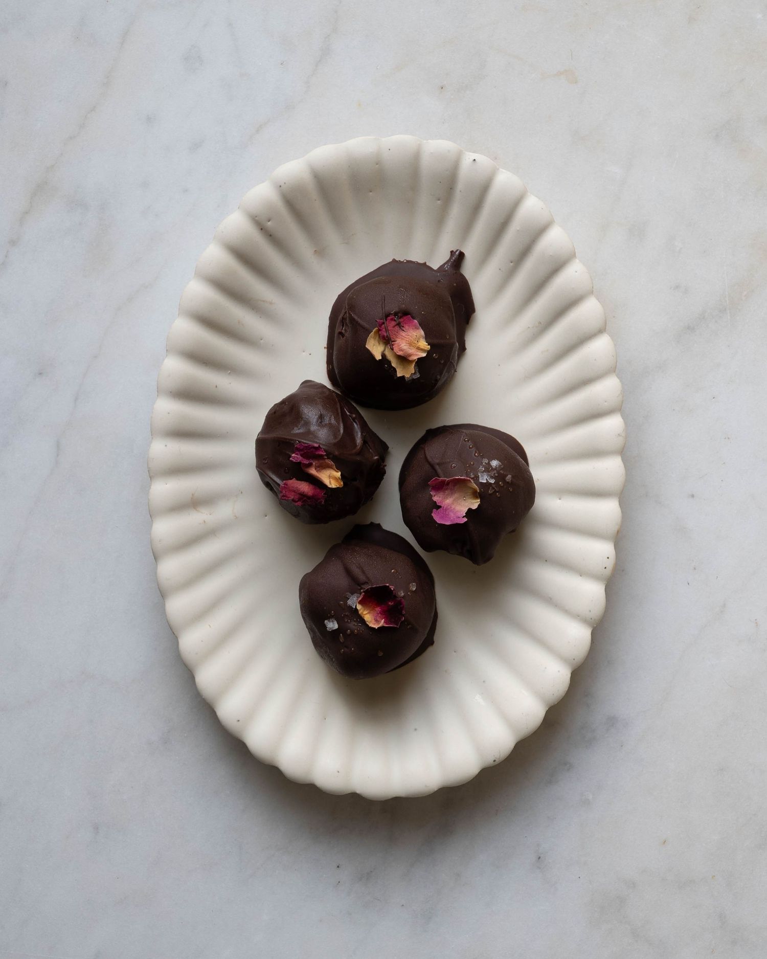 Four dark chocolate truffles decorated with dried rose petals and sea salt on a white fluted plate.