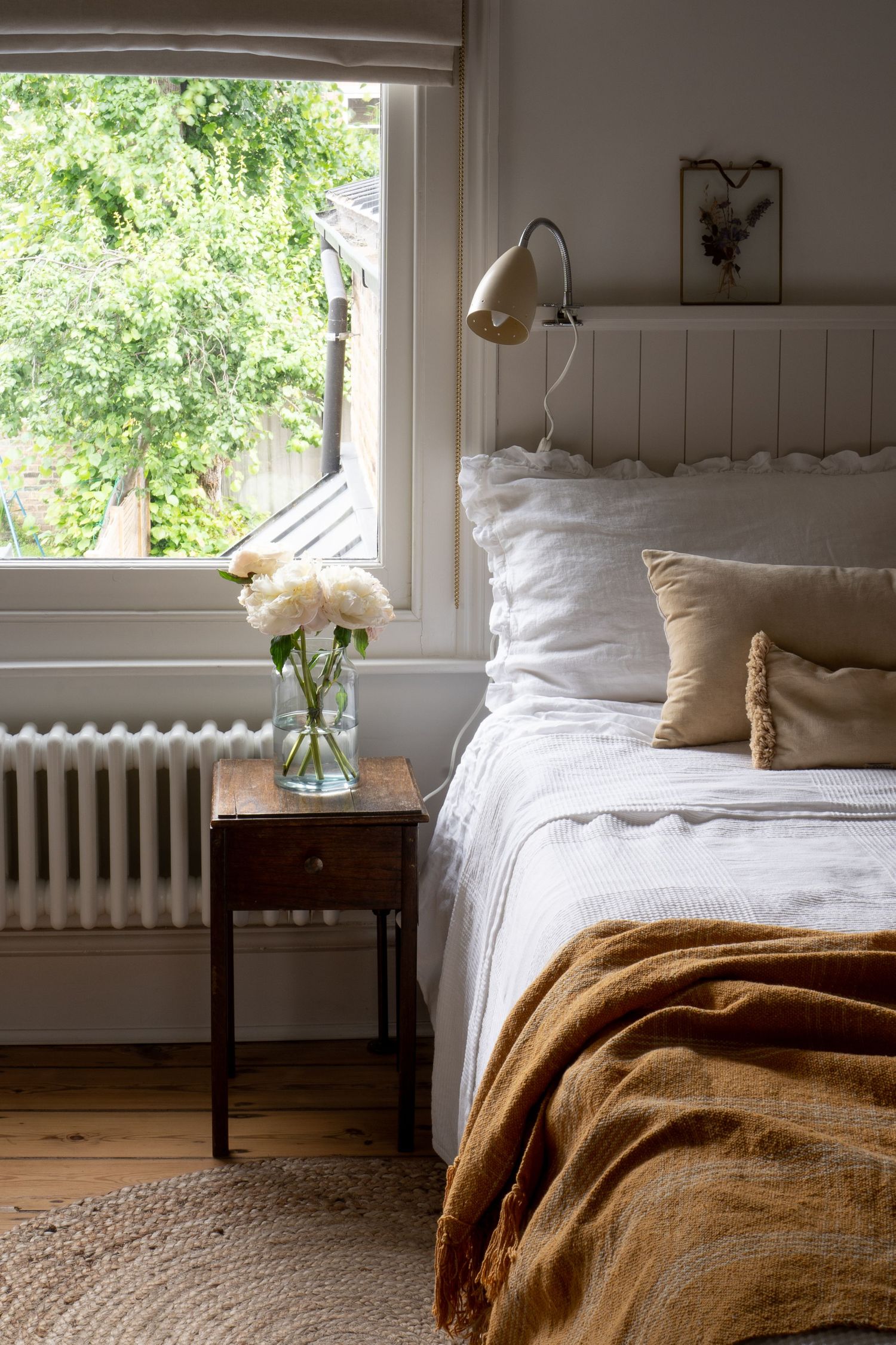 Cozy bedroom with white linen bedding, mustard throw blanket, wooden nightstand with white roses, and garden window view.
