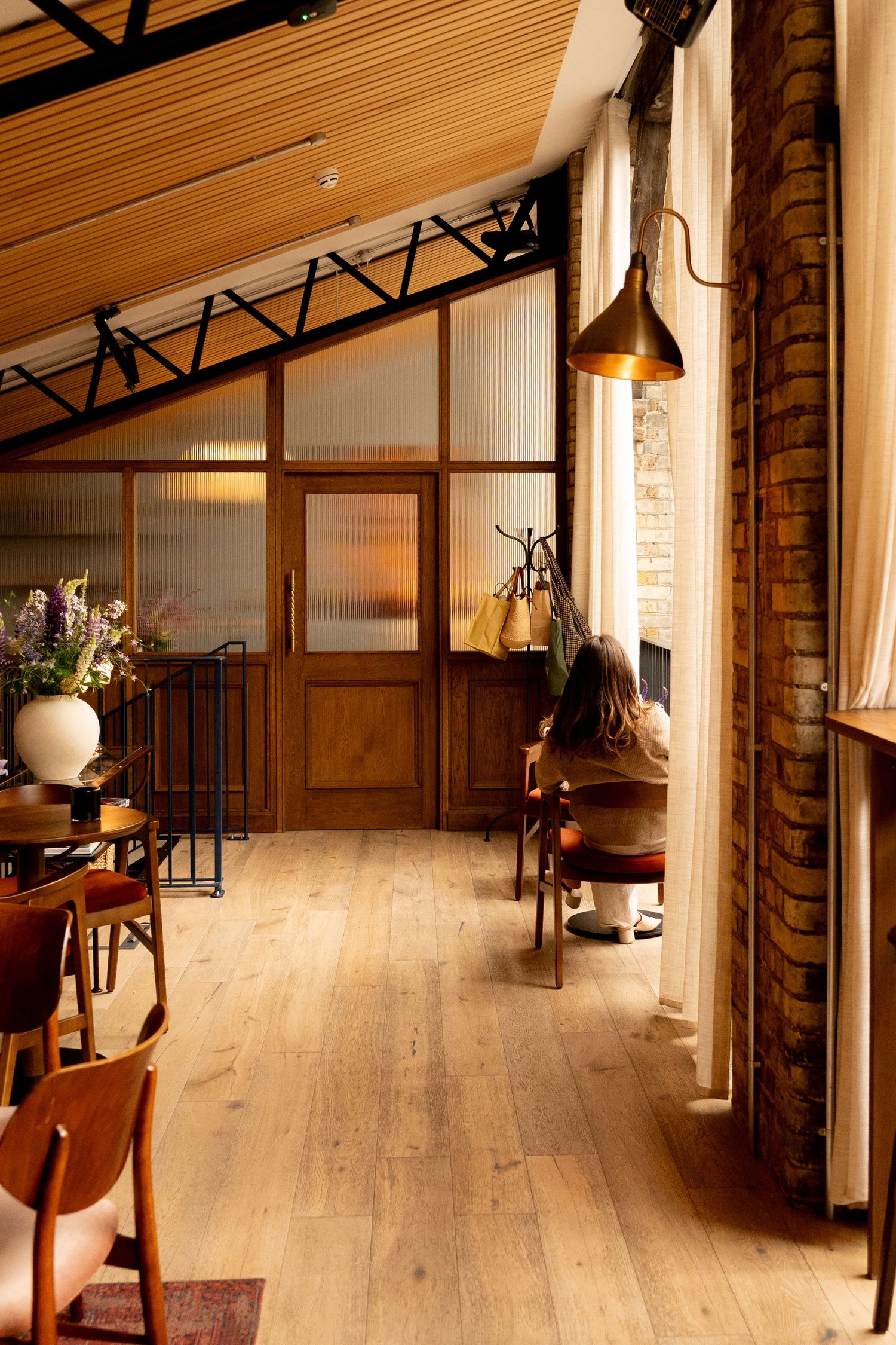 Warm rustic restaurant interior with wooden floors, brick walls, vaulted ceiling, and natural light streaming through tall windows.