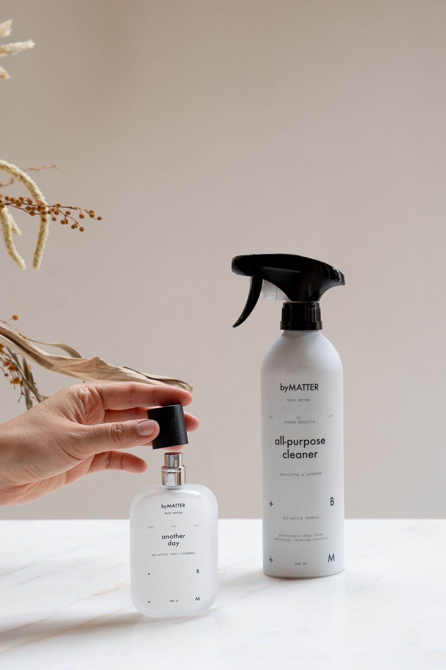 Hand holding byMATTER another day perfume beside all-purpose cleaner spray bottle on marble surface.