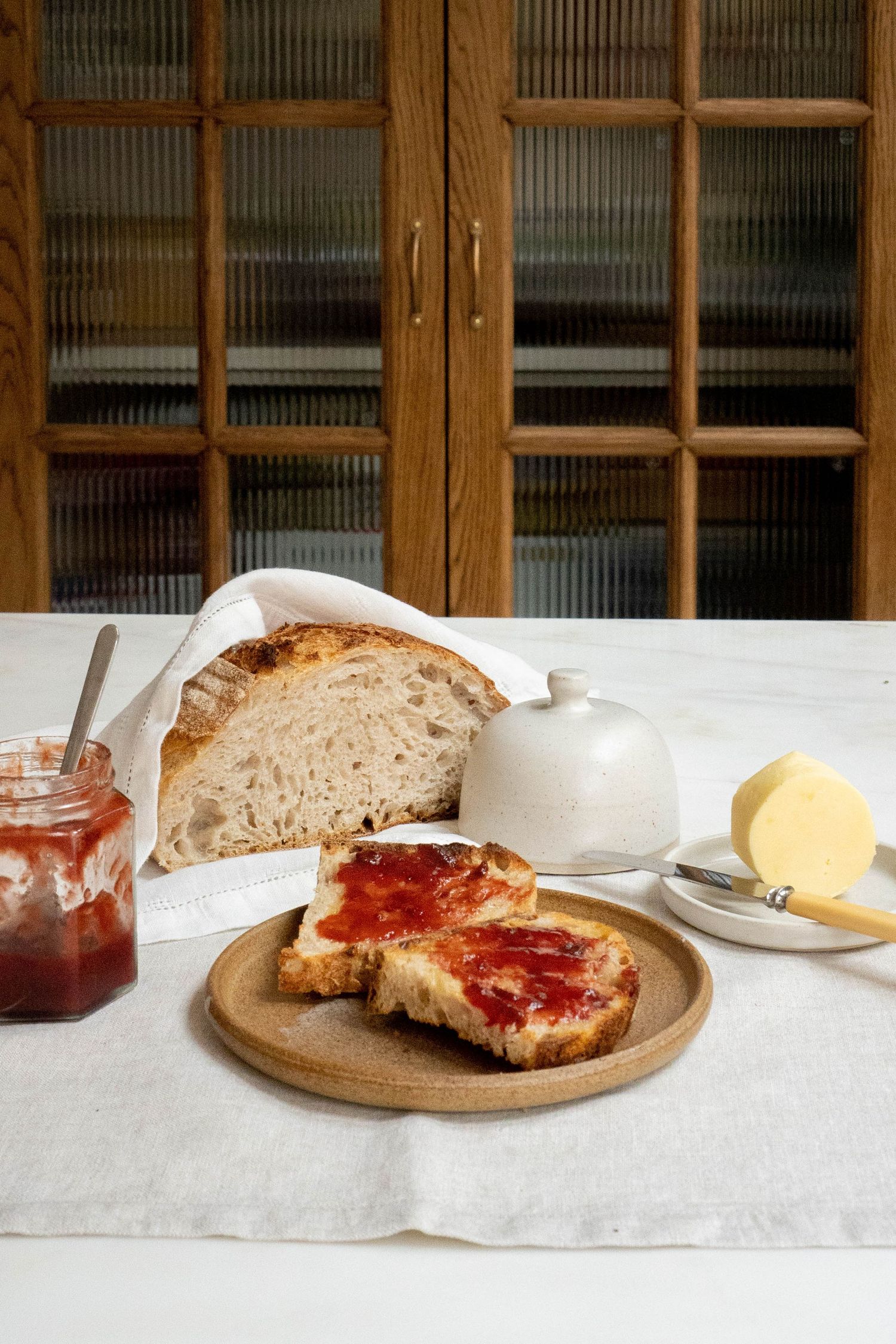 Rustic sourdough bread with strawberry jam on wooden board, butter dish, jam jar on white linen tablecloth.