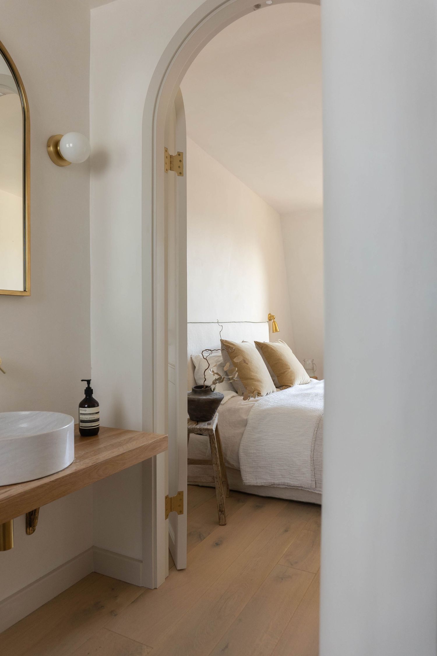 Soft neutral ensuite with arched doorway connecting bathroom vanity to cozy bedroom with gold accents.