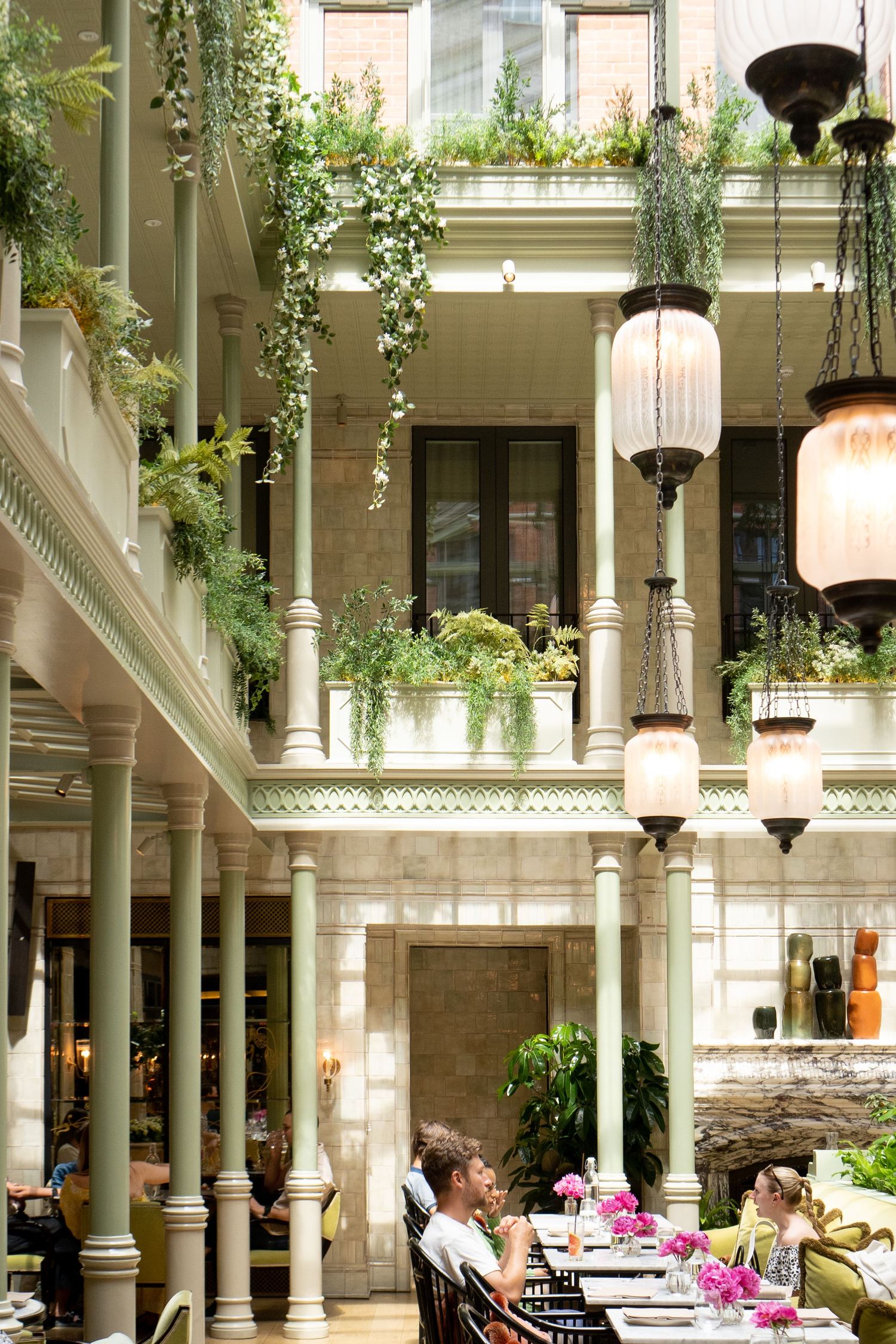 Stunning restaurant atrium with green columns, hanging lanterns, cascading plants, and diners at pink flower-adorned tables.