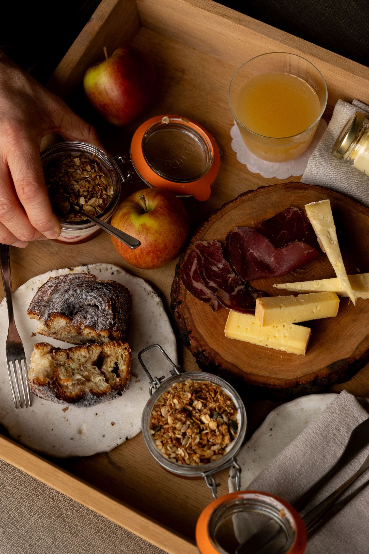Overhead view of artisan breakfast tray with cheese board, sliced bread, fresh apples, granola jars, and apple juice on wooden tray.