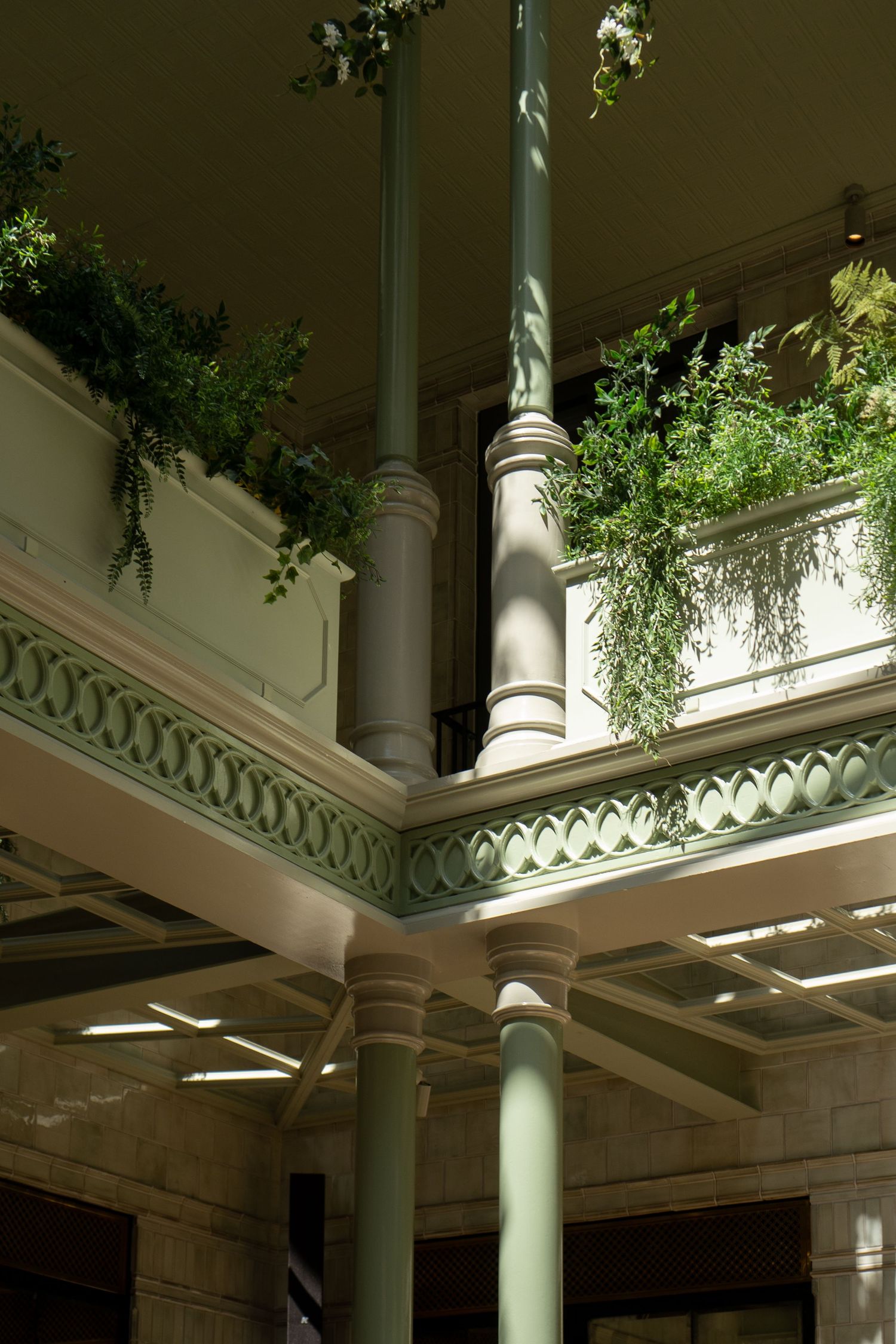 Grand interior atrium with sage green columns, ornate railings, and lush trailing plants in natural light.