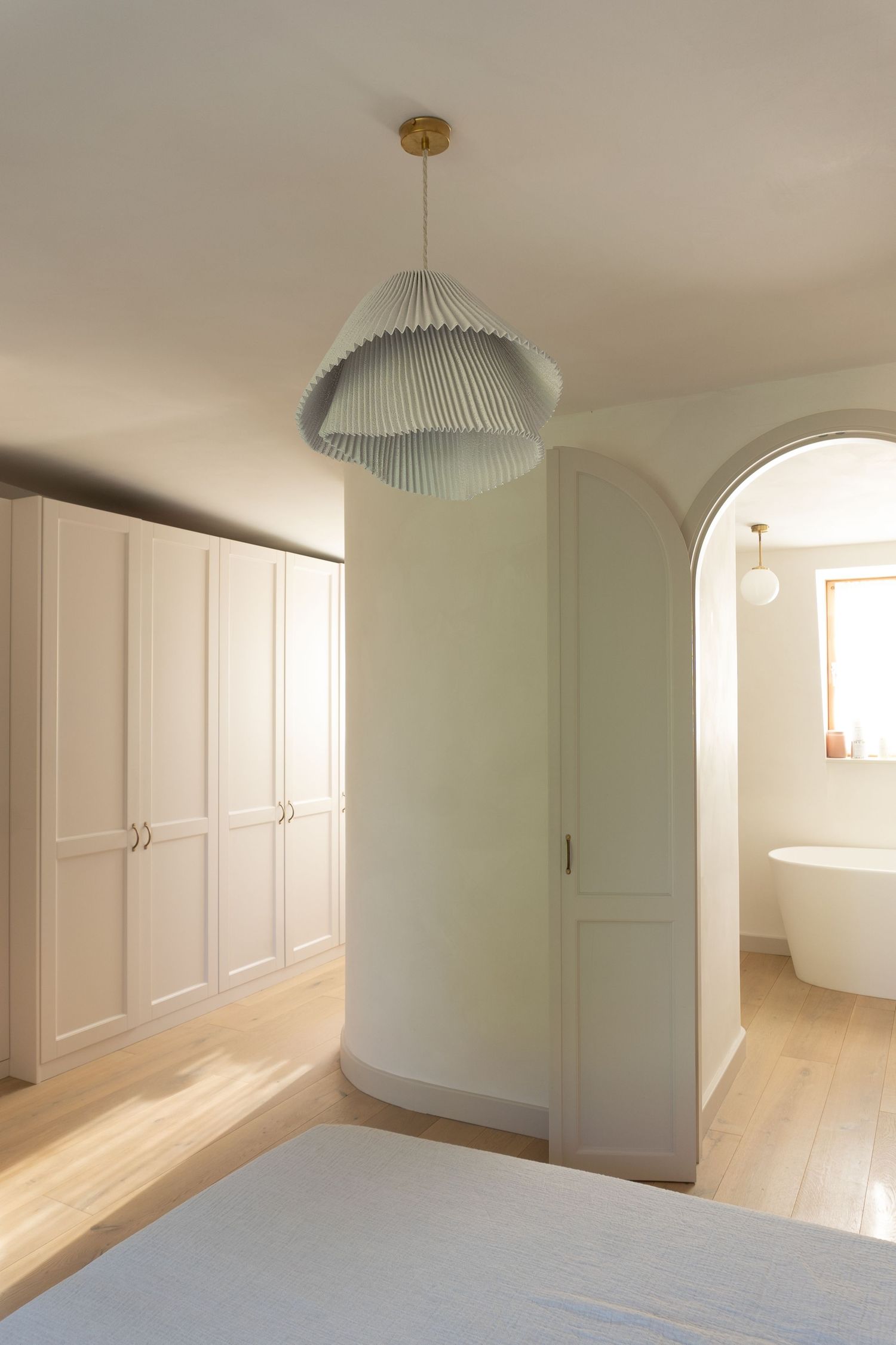 Bright bedroom with built-in wardrobe, pleated pendant light, arched doorway leading to freestanding bathtub.