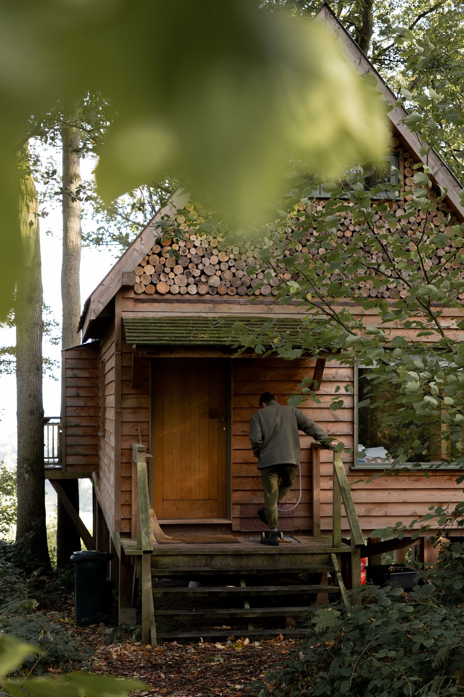 Man entering rustic wooden cabin with log-stacked roof gable, surrounded by dense green forest foliage.
