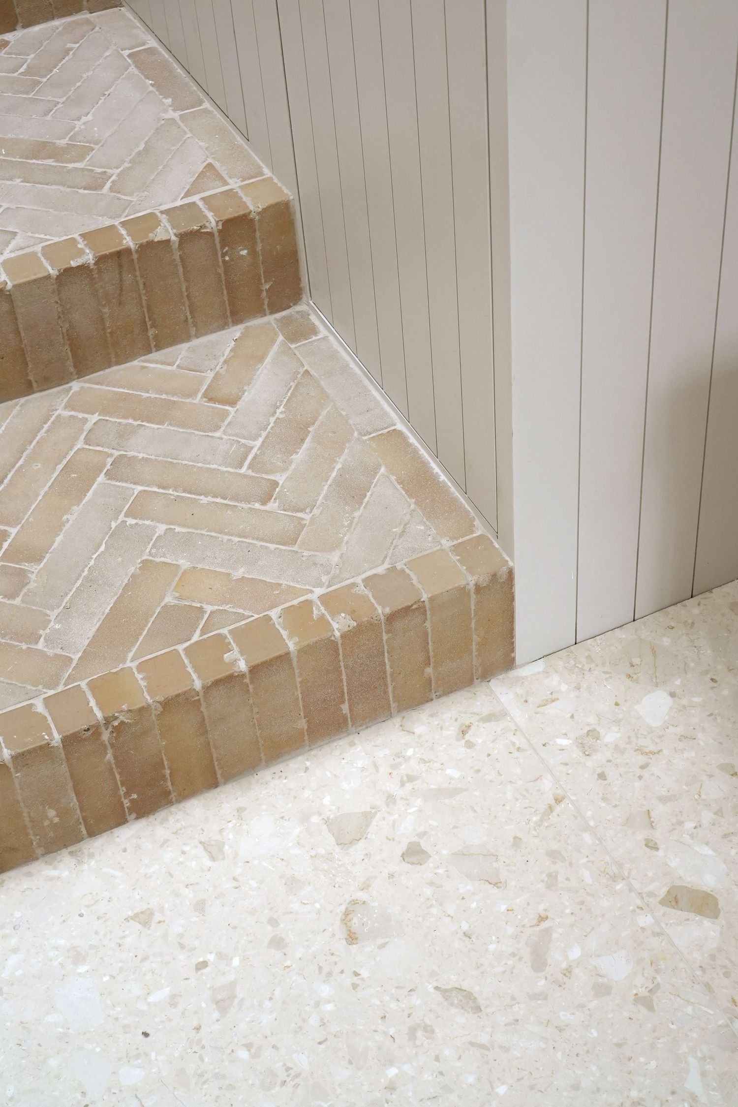 Close-up of herringbone patterned stone stair treads beside white beadboard paneling and terrazzo floor.