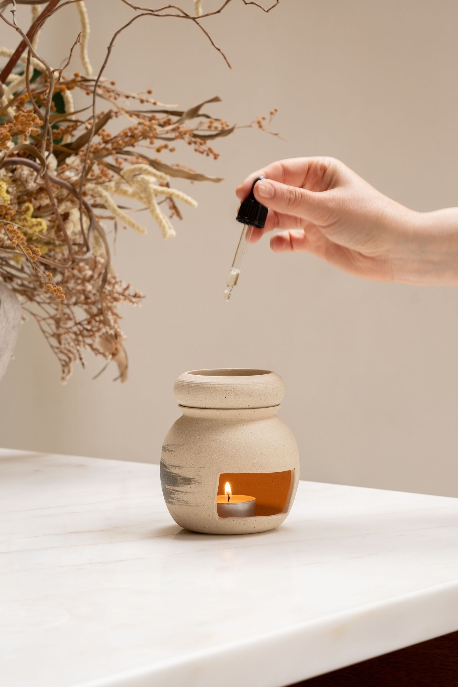 Hand dropping essential oil into a ceramic oil burner with lit candle on a marble surface.